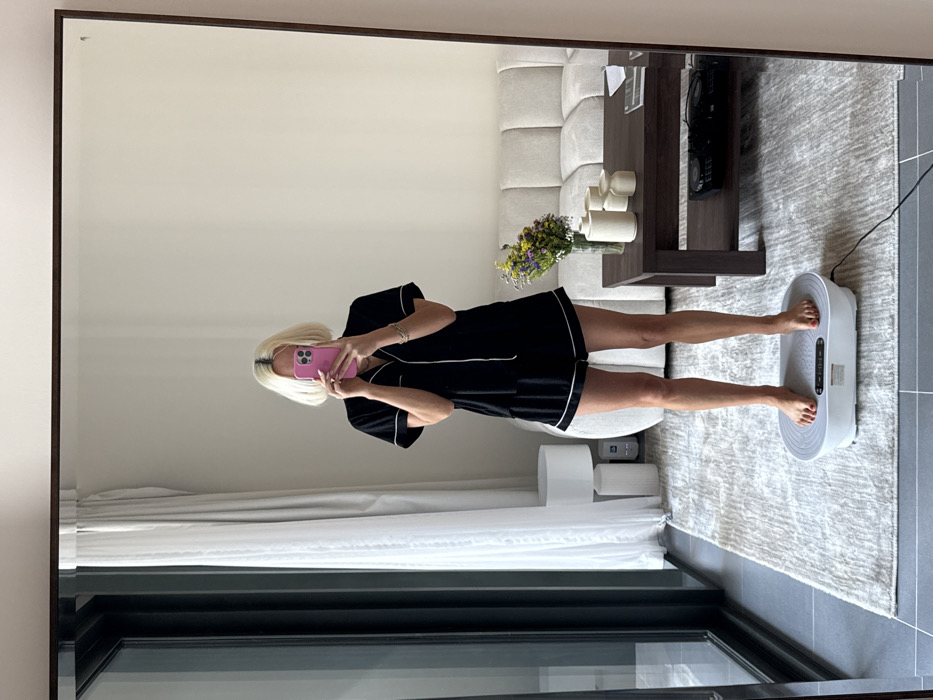 A woman stands on a white vibration plate in her living room, capturing her reflection in a large mirror. She is wearing black pajamas with white piping and holding a pink smartphone to take the picture. The living room features a neutral color palette, with a light gray rug, beige sofa, and dark wood coffee table adorned with white vases and flowers. Curtains are visible in the lower-left corner, and a window is below them.