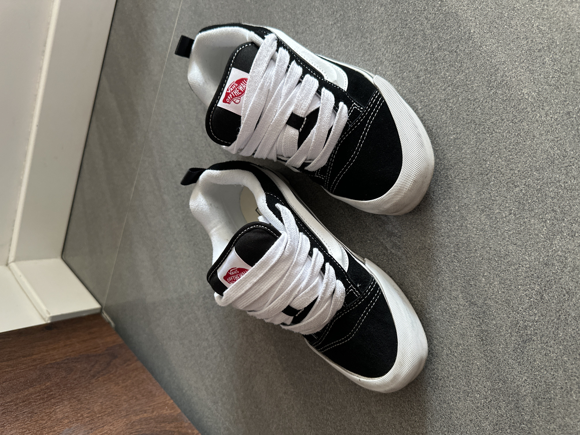A pair of black and white Vans shoes are displayed on a gray tiled floor, situated near a white baseboard and a wooden surface. The shoes feature white laces and the iconic Vans "Off The Wall" logo on the tongue. The lighting suggests an indoor environment, possibly a residential area or a retail space. There is no indication of people or any actions taking place. The image is a still, neutral shot of the footwear. The specific location in Warsaw, Poland, cannot be determined from the visual information provided.