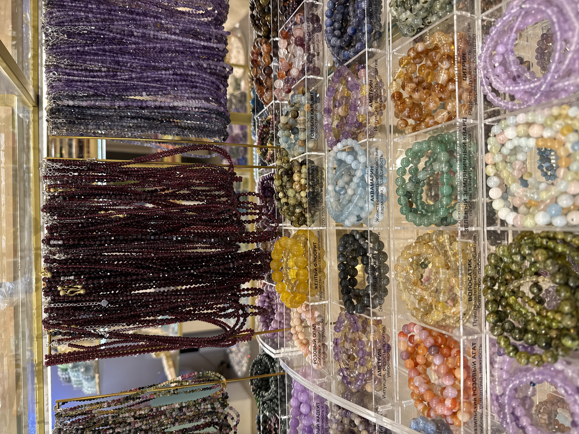 This is a close-up, eye-level shot of a retail display featuring a large assortment of beaded bracelets. The bracelets are predominantly made of colorful gemstones and are organized in clear plastic dividers, creating a grid-like presentation. The variety of colors includes deep purples, reds, oranges, yellows, greens, blues, and pinks. Some bracelets are displayed hanging in dense strands, while others are neatly coiled within their compartments.
Visible text in Russian identifies some of the gemstones and their purported properties. Examples include "РОЗОВЫЙ ОПАЛ" (Pink Opal) associated with love and protection, "ЖЕЛТЫЙ ФЛЮОРИТ" (Yellow Fluorite) linked to creativity and intuition, "АКВАМАРИН" (Aquamarine) described as promoting femininity and calmness, and "ЦИТРИН" (Citrine) associated with joy and wealth. The setting appears to be a jewelry store or a market stall, with reflections visible in some of the display materials, suggesting a well-lit indoor environment. The overall impression is one of abundance and variety in gemstone jewelry.