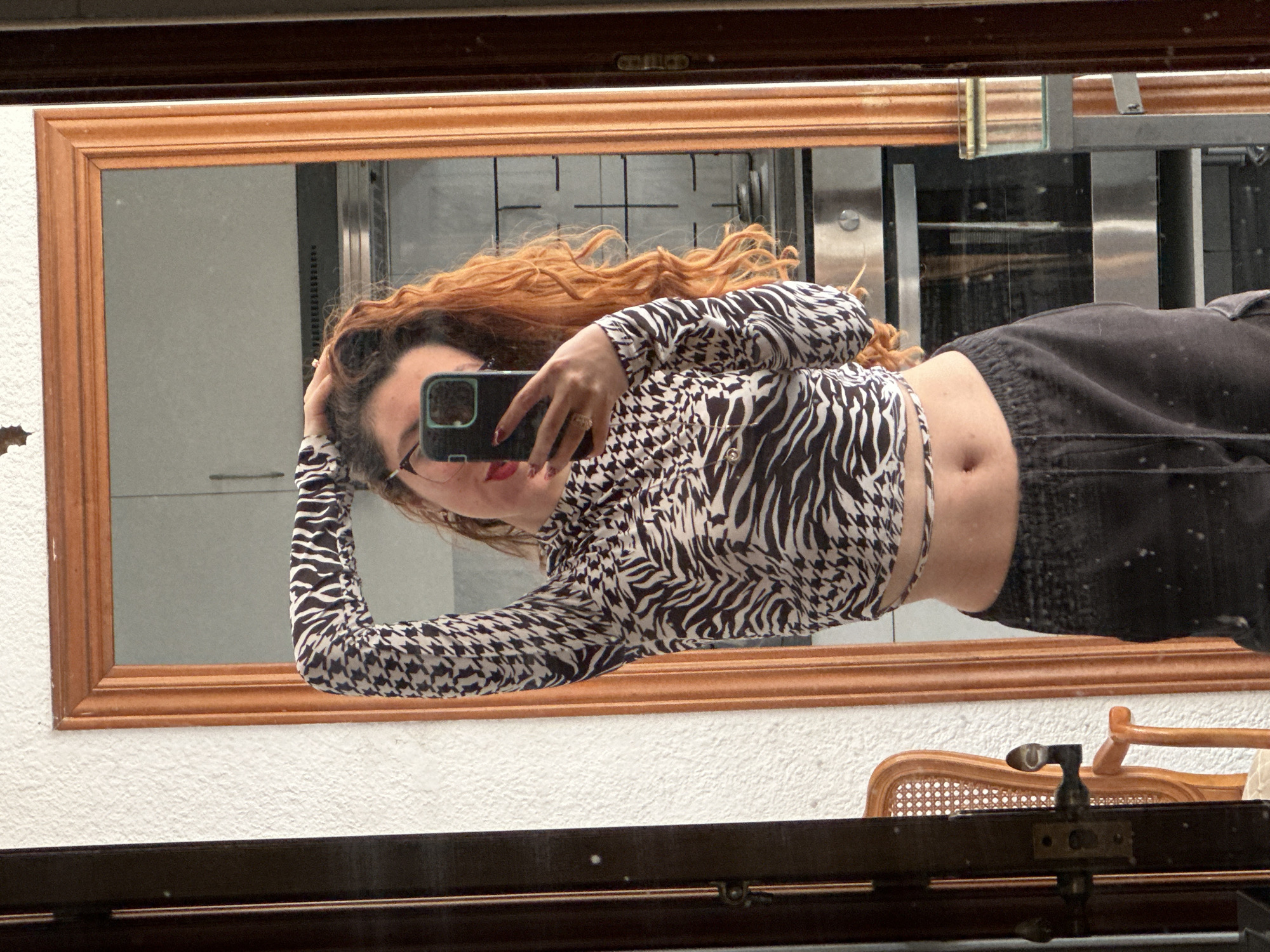 The image shows a person reflected in a mirror. The person has red curly hair, is wearing glasses, a patterned long-sleeved crop top, and dark pants. They are holding a cell phone with a dark case, presumably taking a picture of themselves in the mirror. The background reflected in the mirror appears to be a modern interior with stainless steel elements. The mirror has a wooden frame and is hanging on a white textured wall. Below the mirror, there is a wooden object or piece of furniture with a lighter-colored woven section visible. The reflection suggests an indoor environment, possibly a private space. It is impossible to confirm that the location is in the city of Barcelona, Spain. The image lacks discernible geographical features that would pinpoint such a location.