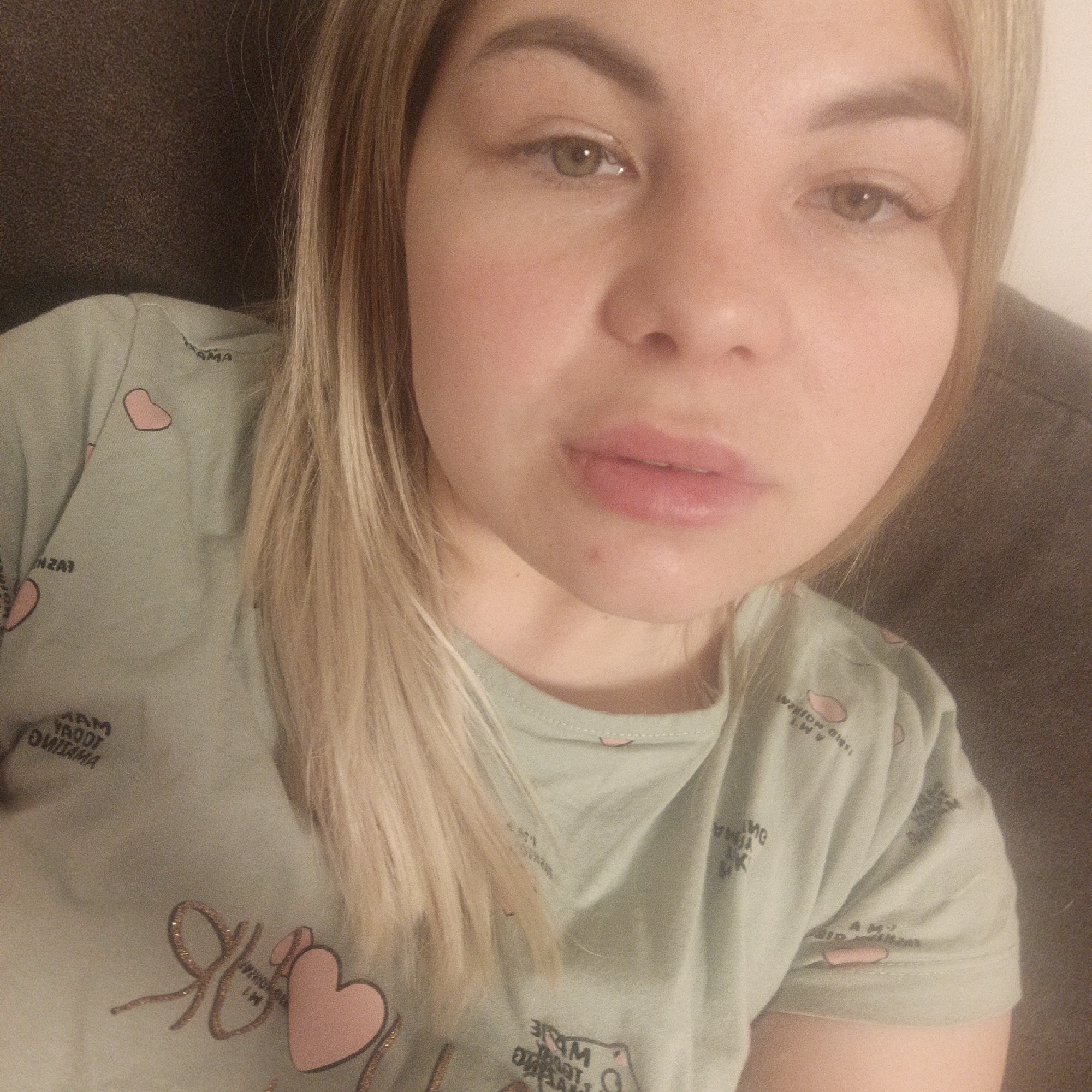A light-skinned woman with long, straight blonde hair and green/hazel eyes is captured in a close-up selfie. Her expression is calm and neutral, featuring full lips and well-defined eyebrows. A small red blemish or mark is visible below her lower lip. Her hair, parted on the side, falls over her shoulders and chest.
She is wearing a light green t-shirt decorated with multiple small pink heart shapes and scattered text. A prominent design on the shirt includes a glittery, cursive "Я" next to a larger pink heart. Various mirrored words and phrases are visible on the fabric; for example, "AMA" is discernible on her right shoulder (likely part of a word like "MAMA"). Other mirrored text includes what appears to be "MAMA YADOT OMA" and other short phrases containing letters such as "AM" or "IM".
The woman is indoors, with a dark, textured background, possibly a couch or upholstered headboard. The lighting suggests an indoor setting, potentially in the evening or at night. The image focuses on her face and upper body, taken from a slightly elevated angle.