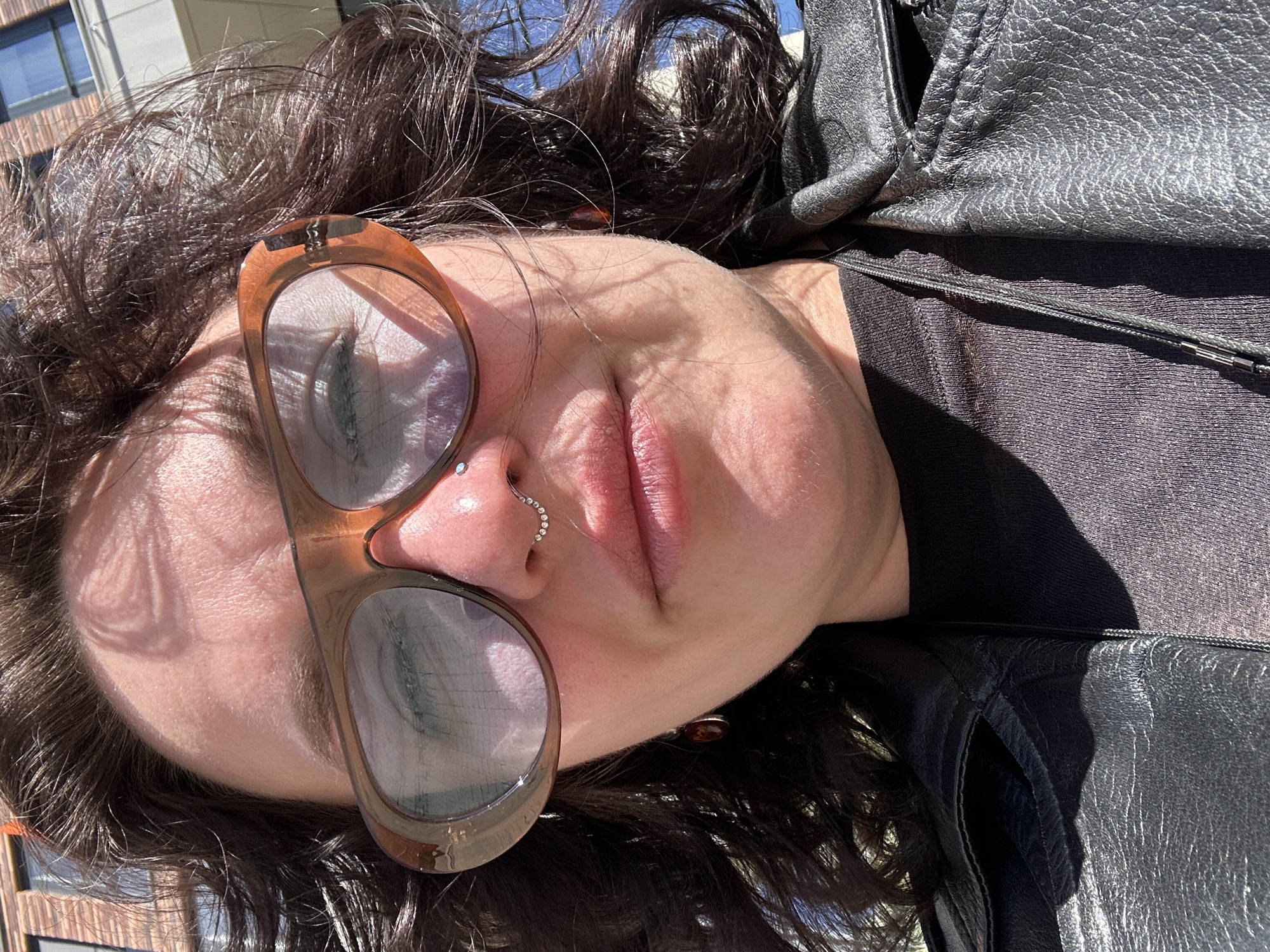 A woman is shown in a close-up, angled portrait. She is wearing glasses with a brown frame, and a nose ring with multiple stones. She is wearing a black shirt and a black leather jacket. Dark brown hair frames her face. The background shows a building exterior and a patch of blue sky. The image does not provide information to establish the location as Izhevsk, Russia.