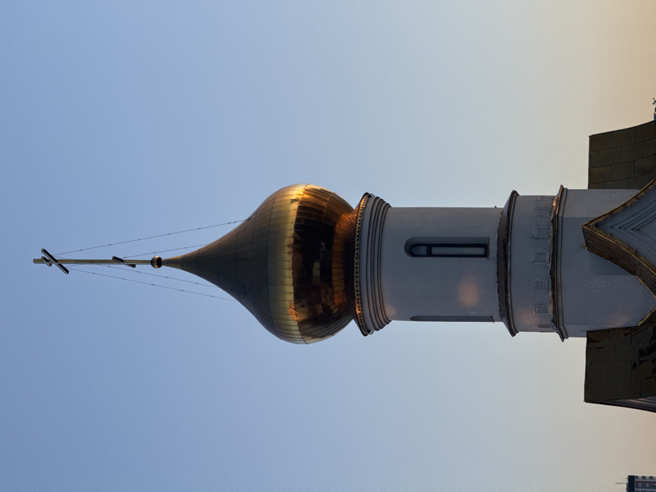 A golden, onion-shaped dome of an Orthodox church bell tower rises against a clear, light blue sky. The dome, reflecting warm light, is topped by a dark Orthodox cross with three horizontal bars. Thin wires or rods stabilize the cross, connecting it to the dome's apex. The dome rests on a white, cylindrical drum that features a single, tall, narrow, arched window with a dark frame. Below this drum, the bell tower extends downward as a wider, white cylindrical section, displaying horizontal banding and small rectangular vents. A portion of a golden-colored roof with a pointed gable is visible at the base of the tower structure within the frame. The scene depicts a detail of the Church of the Icon of the Mother of God, "The Inexhaustible Chalice", located in the Bibirevo District of Moscow, Russia.
