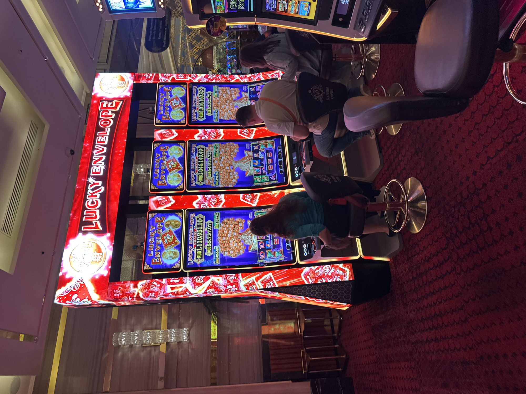 The image depicts a section of a casino floor in Estosadok, Russia, featuring several "Lucky Envelope" slot machines. The machines are brightly lit with red and gold accents, displaying graphics of envelopes and coins. Three people are visible playing the machines: two women and one man. The women are seated on bar stools facing the machines, while the man is also seated and actively engaged with his machine. One of the women is wearing a teal top, and the man is wearing a light-colored shirt. The casino floor is carpeted with a red pattern. In the background, other slot machines and casino elements are visible, including a portion of a sign that appears to read "KASHO COCH". The overall environment suggests a lively and engaging casino setting.