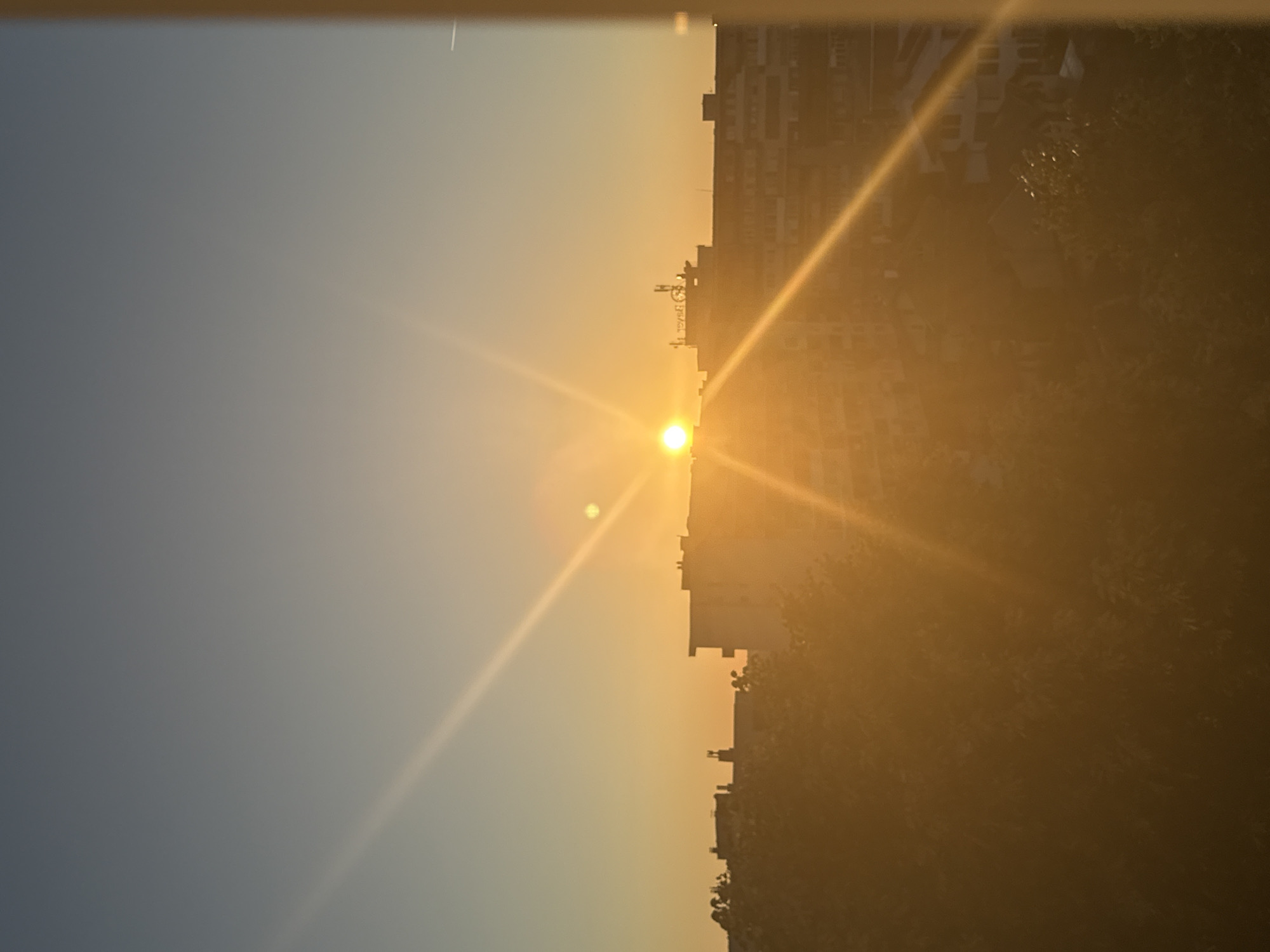 This is a vertical, eye-level, medium shot of a sunset over the city of Dobrich, Bulgaria. The sun is low on the horizon, casting a warm, golden light across the sky and silhouetting buildings and trees. The sky transitions from a clear blue at the top to a soft orange near the horizon. Sunlight flares are visible, emanating from the sun in various directions. In the mid-ground, a line of buildings, appearing to be apartment blocks, is visible, with some trees in the foreground obscuring the lower portions of the structures. The buildings have flat roofs and some appear to have antennae or other rooftop structures. The overall impression is one of a tranquil urban sunset.