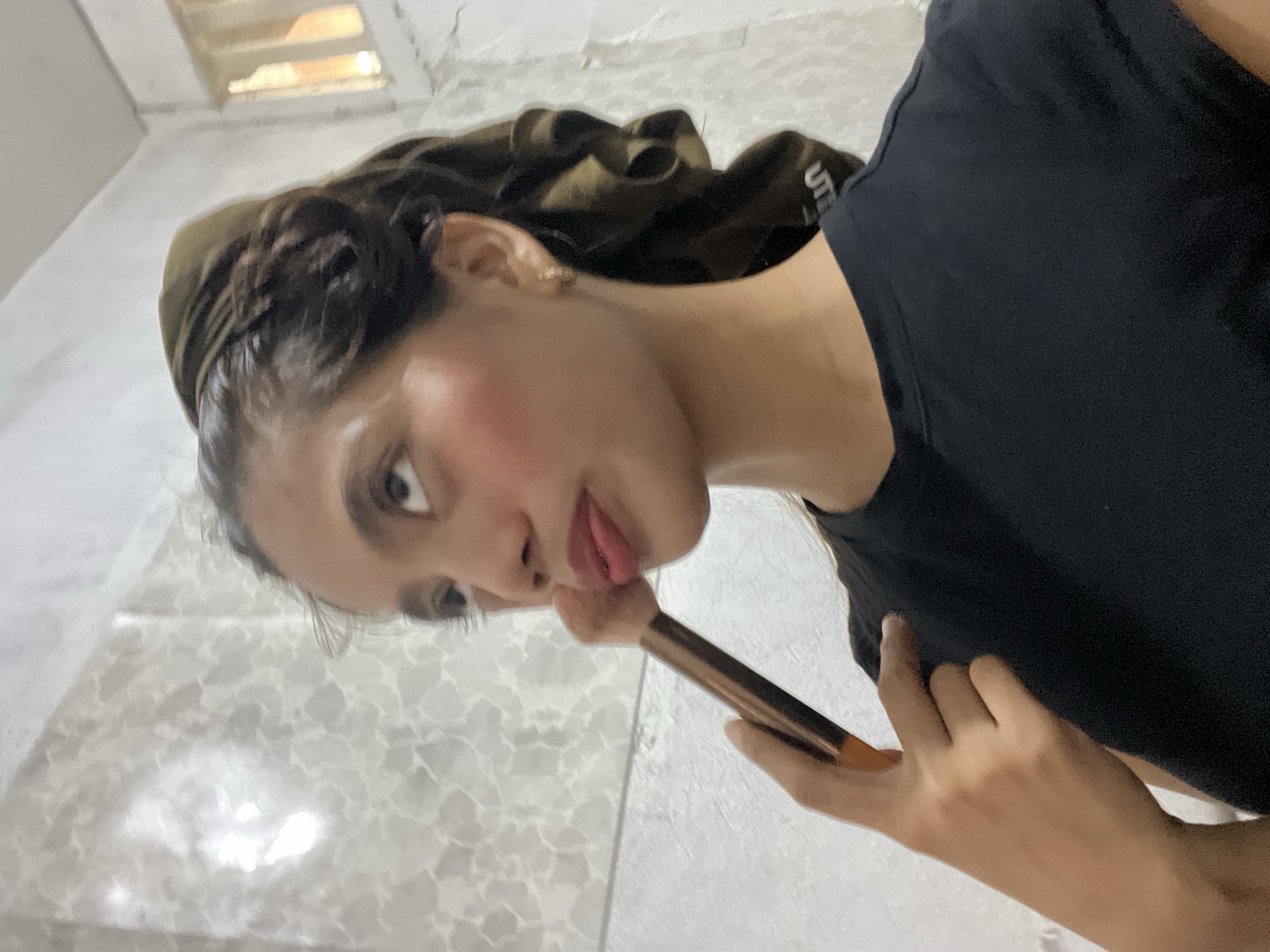 A tilted, close-up indoor portrait features a young woman, likely in Davao City, Philippines, looking down playfully at the camera. Her tongue is slightly extended, conveying a candid and lighthearted mood. She holds a makeup brush with a rose gold and black handle near her right cheek, where a subtle application of blush is visible. Her dark hair is pulled back, appearing to be partially covered by a dark olive-green fabric or scarf. She wears a plain black t-shirt, and the letters "UT" are visible on the fabric near her left shoulder, suggesting a Uniqlo UT brand. The background consists of light-colored, possibly tiled walls and floor, indicating a domestic setting. In the upper background, a vent or window allows bright light to enter, hinting at daytime. The image captures a moment of personal care or playful self-expression.