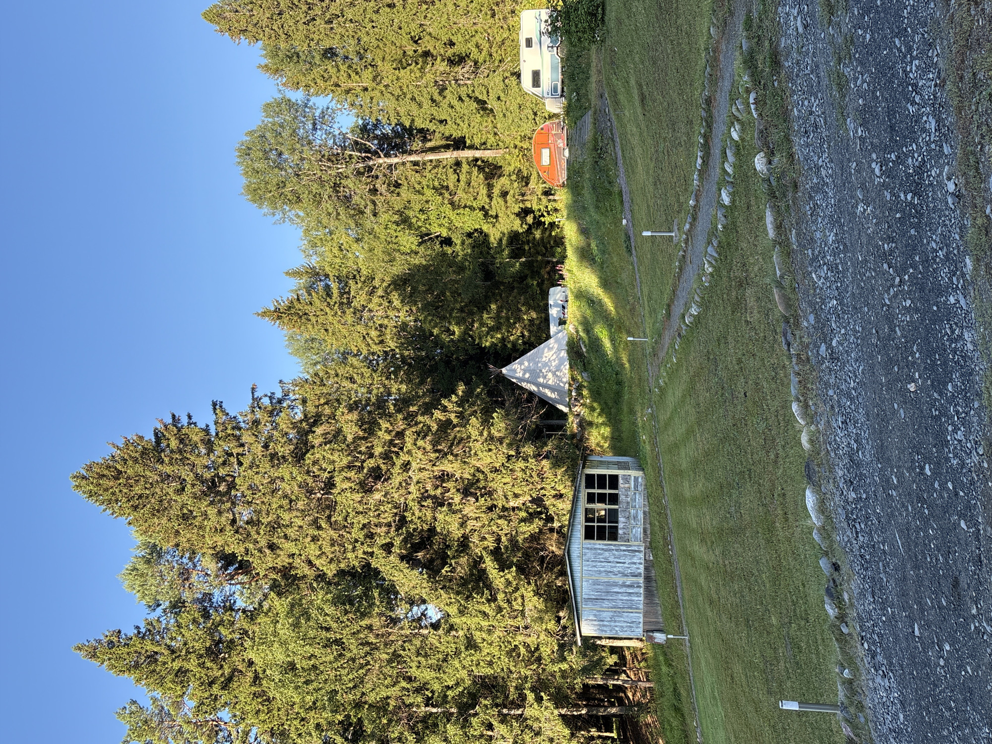 The image depicts a campground scene at dusk in Rautalakhti, Russia. Dominating the background is a dense forest of evergreen trees, casting long shadows on the grassy slopes. In the middle ground, a small, weathered wooden shed with multiple windows stands on a slight incline. To the right of the shed, a white teepee-like tent is visible. Further to the right, a small orange caravan and a white and blue camper van are parked on a grassy area. A winding gravel path, bordered by stones, leads up the slope towards the vehicles. The foreground consists of a gravel-covered area, suggesting a parking or entry point to the campground. The sky is a clear, deep blue, indicative of clear weather at the end of the day. There are no people visible in the image.