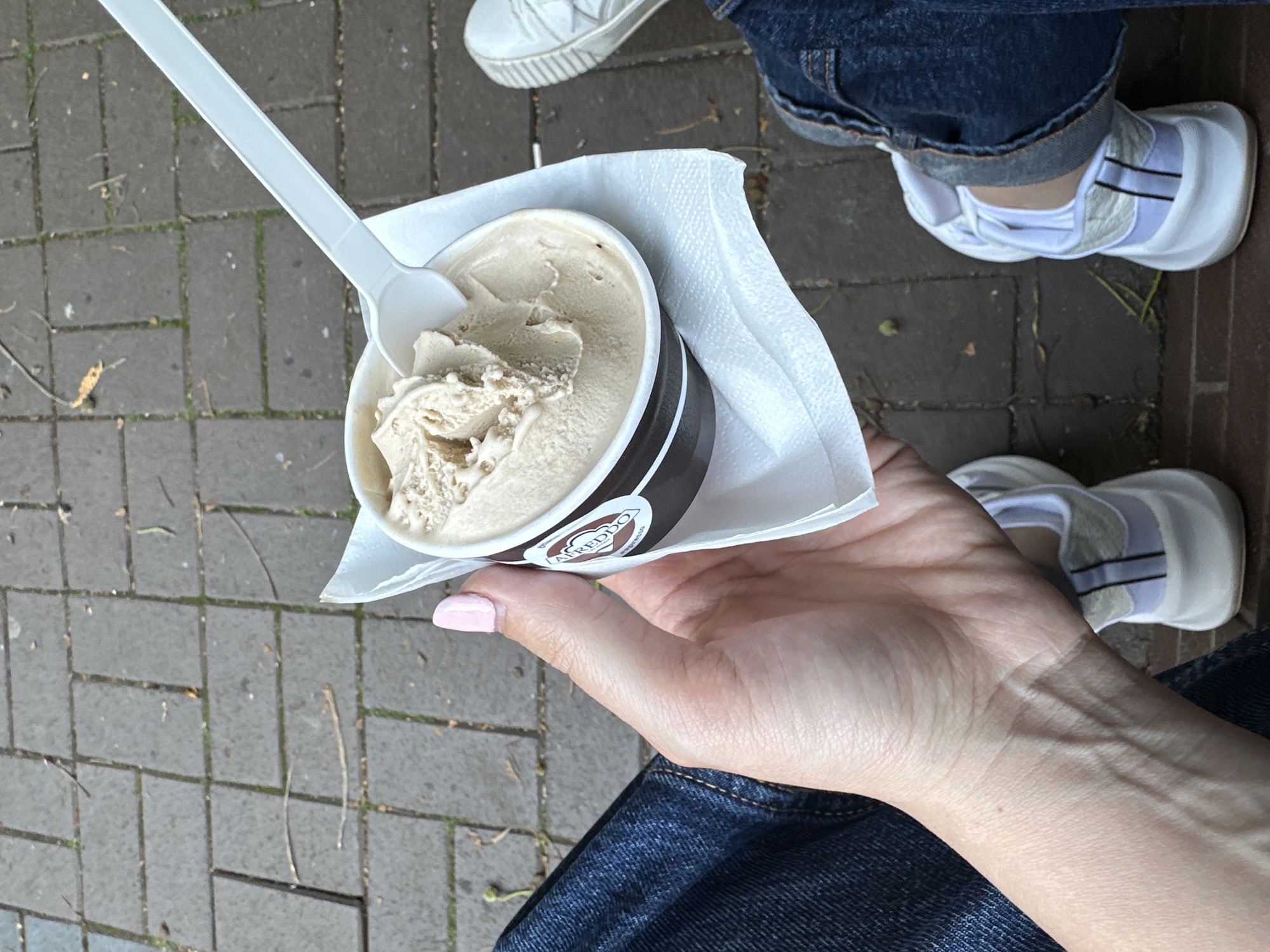 The image shows a first-person perspective of someone holding a cup of ice cream with a white spoon. The cup has a brown band with a logo that appears to say "AMEDDO". The person is wearing blue jeans and a white sneaker is visible to the right. The ground is paved with grey rectangular bricks. The scene takes place outdoors, likely in a public area. There are no visible landmarks or clues that would indicate the location is specifically in Sofia, Bulgaria.