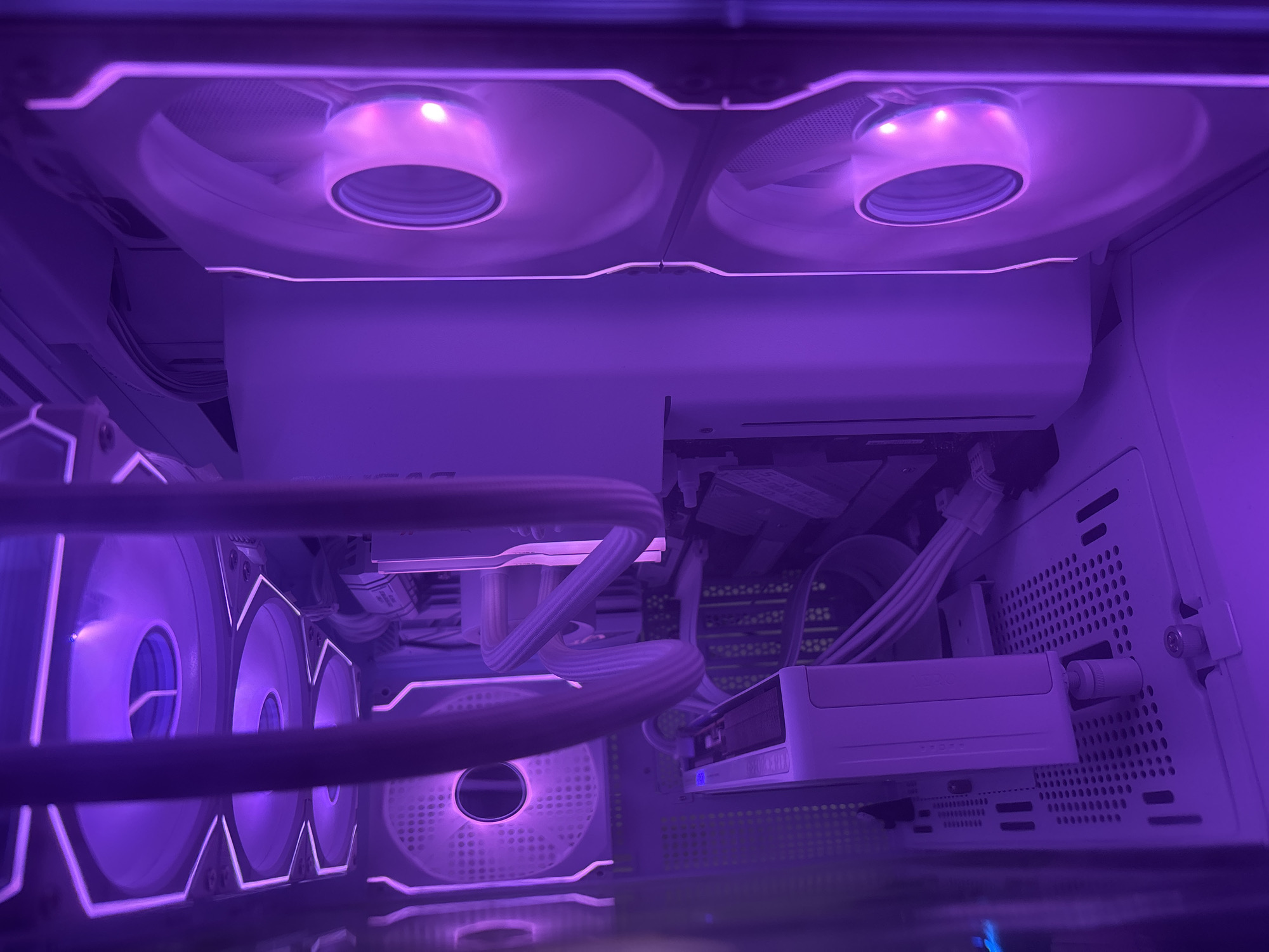 The image shows the interior of a white computer case, illuminated by purple LED lighting. Several large, circular computer fans with white blades and integrated RGB lighting are visible. Some of the fans are mounted on the top of the case, while others are mounted on the side. A white computer component, possibly a graphics card or a CPU cooler block, is also visible, along with white cables connecting various components. The overall impression is of a modern, high-performance computer setup. The location is not discernable from the image.