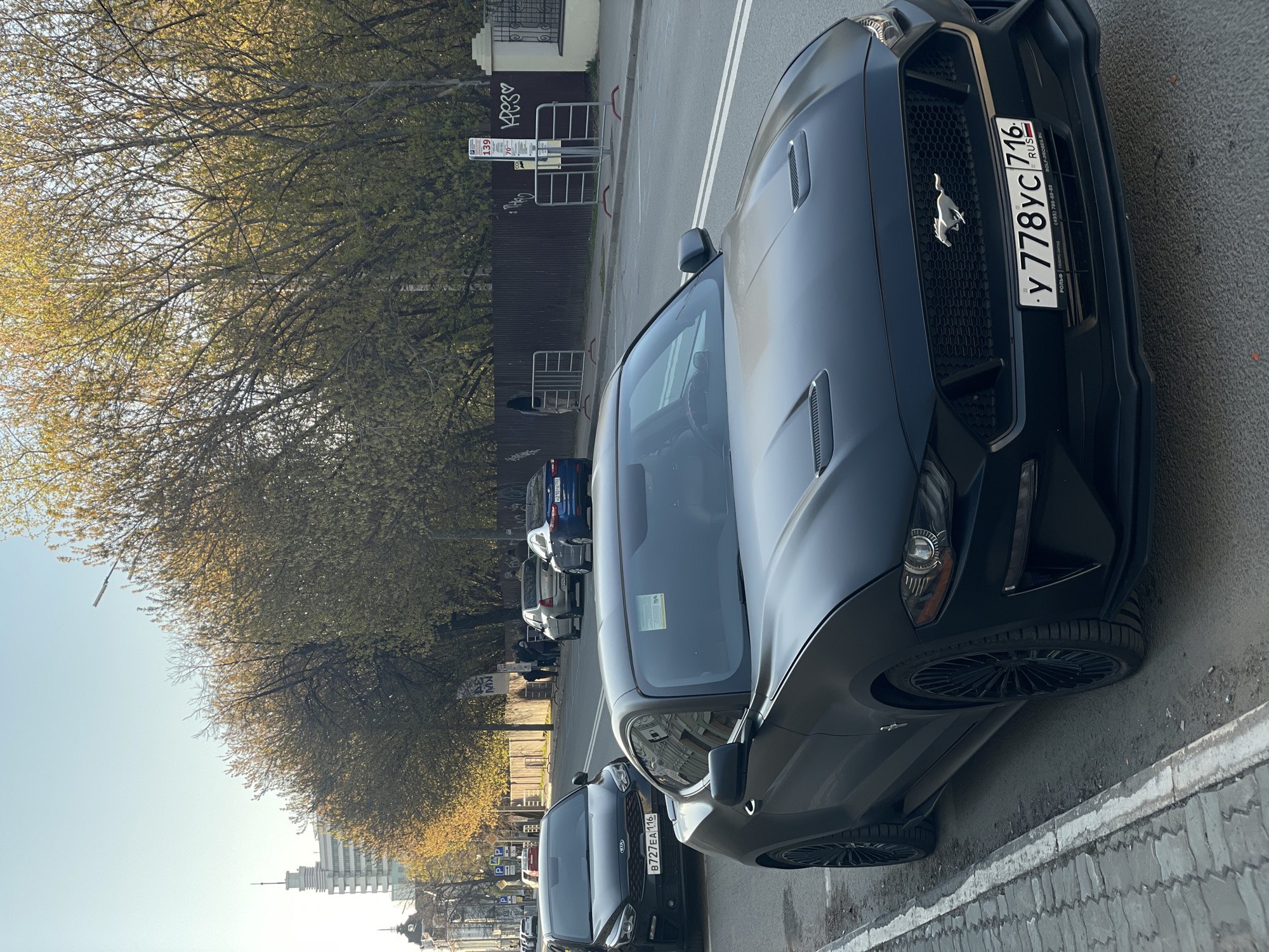 The image captures a street scene in Kazan, Russia, showcasing several parked vehicles along a tree-lined avenue. Dominating the foreground is a black Ford Mustang, its front end prominently displaying the license plate "У 778УС 716" with Russian text indicating "РОЛЬФ | СЕВЕРО-ЗАПАД," a phone number, and "ROLF-PROBEG.RU." The number plate identifies the car's origin as Russia. To the left and slightly behind the Mustang is a black Kia, its license plate reading "B727EA 116." A line of other vehicles stretches further back, including a blue car.
On the left side of the street, a row of trees lines the sidewalk, their branches partially obscuring a building and a bus stop sign marked "139." The sign features a graffiti-style heart and the word "КРЕЗ♡." In the distant background, city buildings and a clear, blue sky are visible, establishing an urban environment with a sunny, daytime setting. The street surface appears to be well-maintained, with white lane markings.