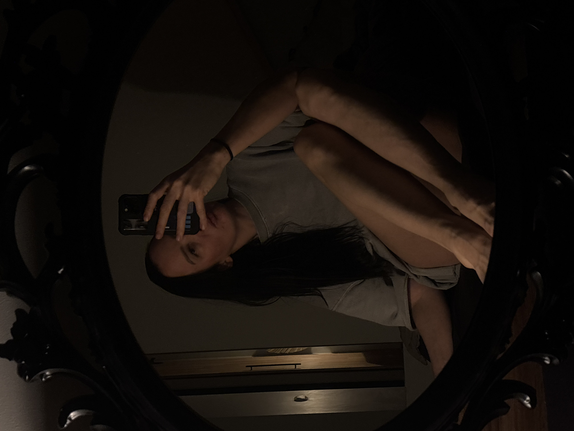 An artistic mirror selfie depicts a person with long dark hair, wearing a light grey long-sleeved top, reflected in an ornate, dark-framed oval mirror. The subject holds a dark smartphone, partially obscuring their face, with a black bracelet visible on one wrist. Their body is positioned with bent legs, creating a somewhat contorted pose within the mirror's reflection. The scene is dimly lit, suggesting evening or night, contributing to a moody and introspective atmosphere. In the background of the reflection, a piece of dark furniture, possibly a dresser, with a handle, and what appears to be a light source or window, are visible. The image captures a quiet, indoor moment, possibly in Chaikovsky, Russia. No discernible text is visible.