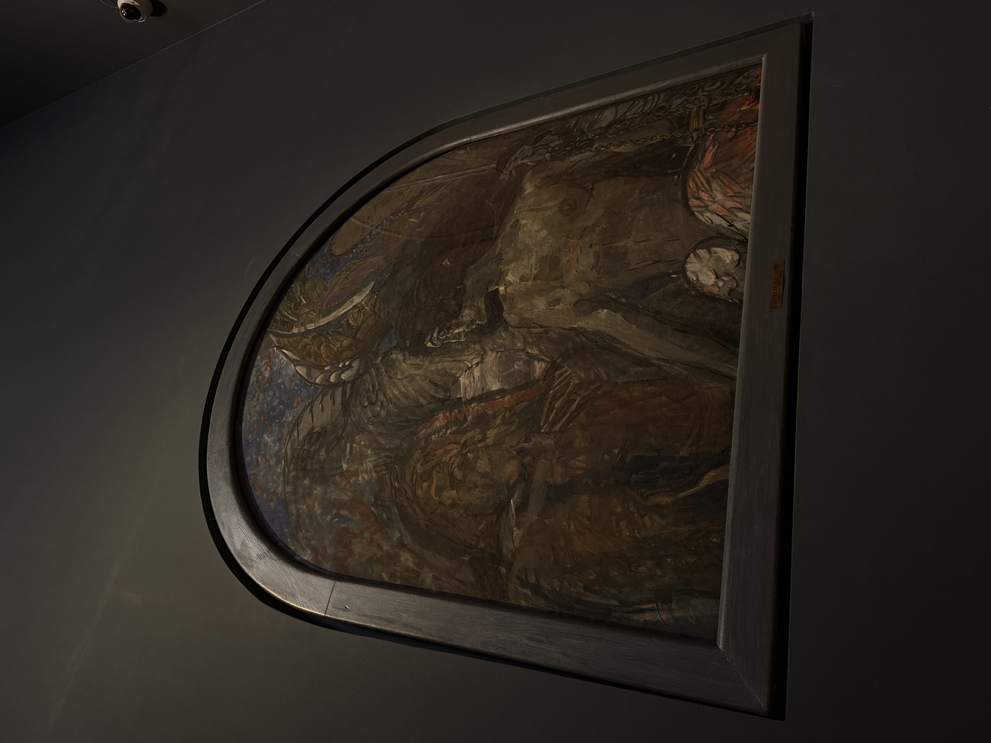 The image shows a painting hanging on a dark gray wall, likely in a museum or gallery. The painting is in a dark, ornate frame that is curved on one side. The painting itself has an abstract, dark color palette of browns, reds, and blues, with the composition appearing chaotic and potentially depicting figures or mythical scenes, though obscured by the painting's style. Above the painting, a security camera is mounted on the wall. Based on the title provided of 'Mikhail Vrubel - The Demon Cast Down - State Tretyakov Gallery - Moscow', the painting is located in the State Tretyakov Gallery in Moscow, Russia.
