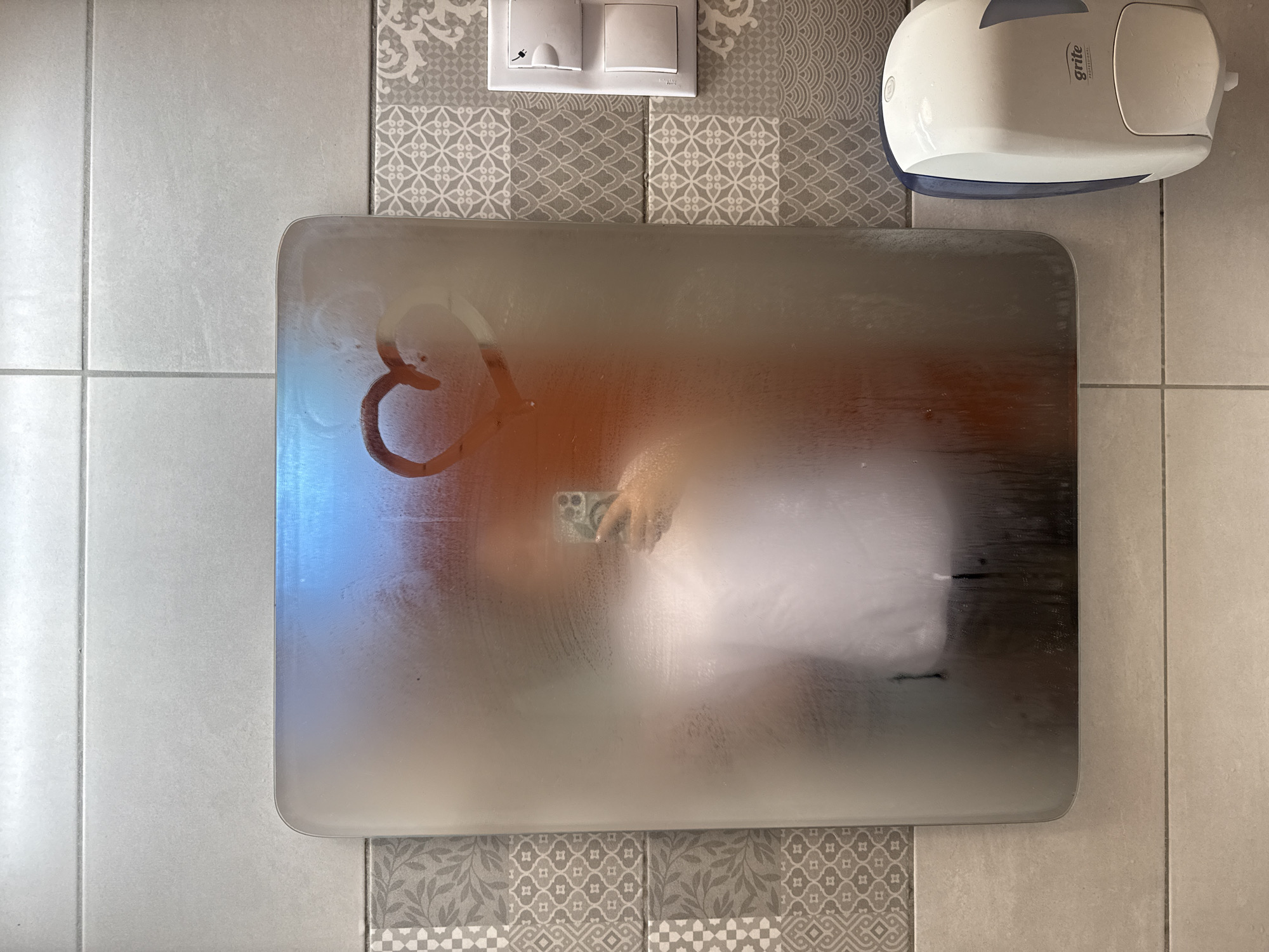 The image shows a close-up, eye-level, interior shot of a bathroom mirror. The mirror is partially fogged, with a heart shape drawn on it. A person's reflection is visible in the mirror, holding a smartphone. To the right of the mirror, there is a white soap dispenser mounted on a tiled wall. The wall tiles are a mix of light gray and patterned gray tiles. The image does not provide any specific indicators of its location within the city of Ventspils, Latvia.