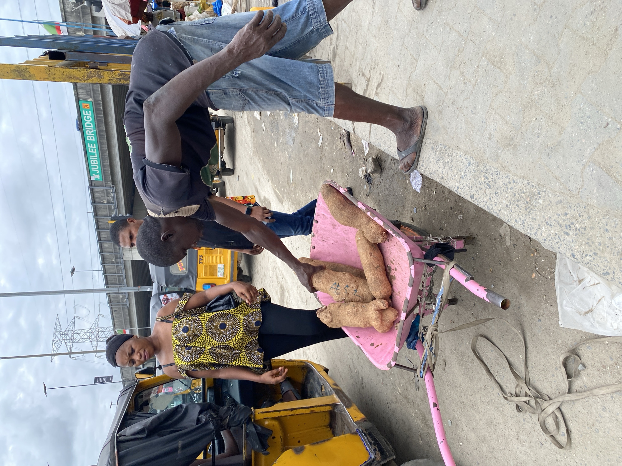 A woman in a yellow patterned top and black pants stands next to a yellow auto-rickshaw (tuk-tuk). A man is handing her yams from a pink wheelbarrow. Another man stands nearby. The scene is on a street with a bridge labeled "JUBILEE BRIDGE" overhead. This suggests the location is Lagos, Nigeria. The ground is concrete with litter. Overhead are electrical cables on poles.