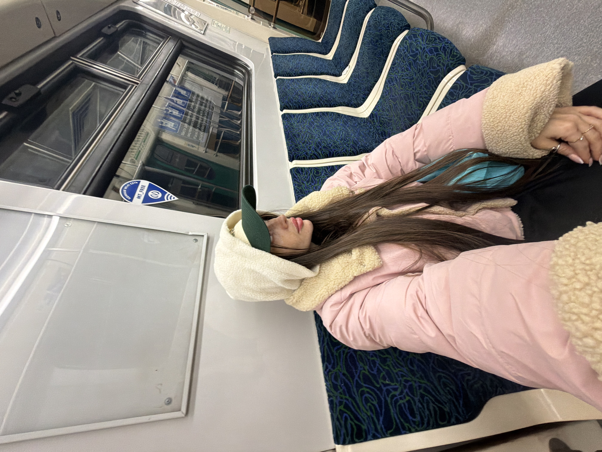 A woman is lying across several vacant seats on a train, possibly a subway car, in Saint Petersburg, Russia. She is wearing a pink jacket with a cream-colored fleece lining and cuffs, a white hooded sweatshirt, and a green baseball cap. She has long, dark brown hair. The seats are upholstered in a dark blue fabric with a patterned design. A window with informational signage is visible to her left. The interior of the train car is white and grey.