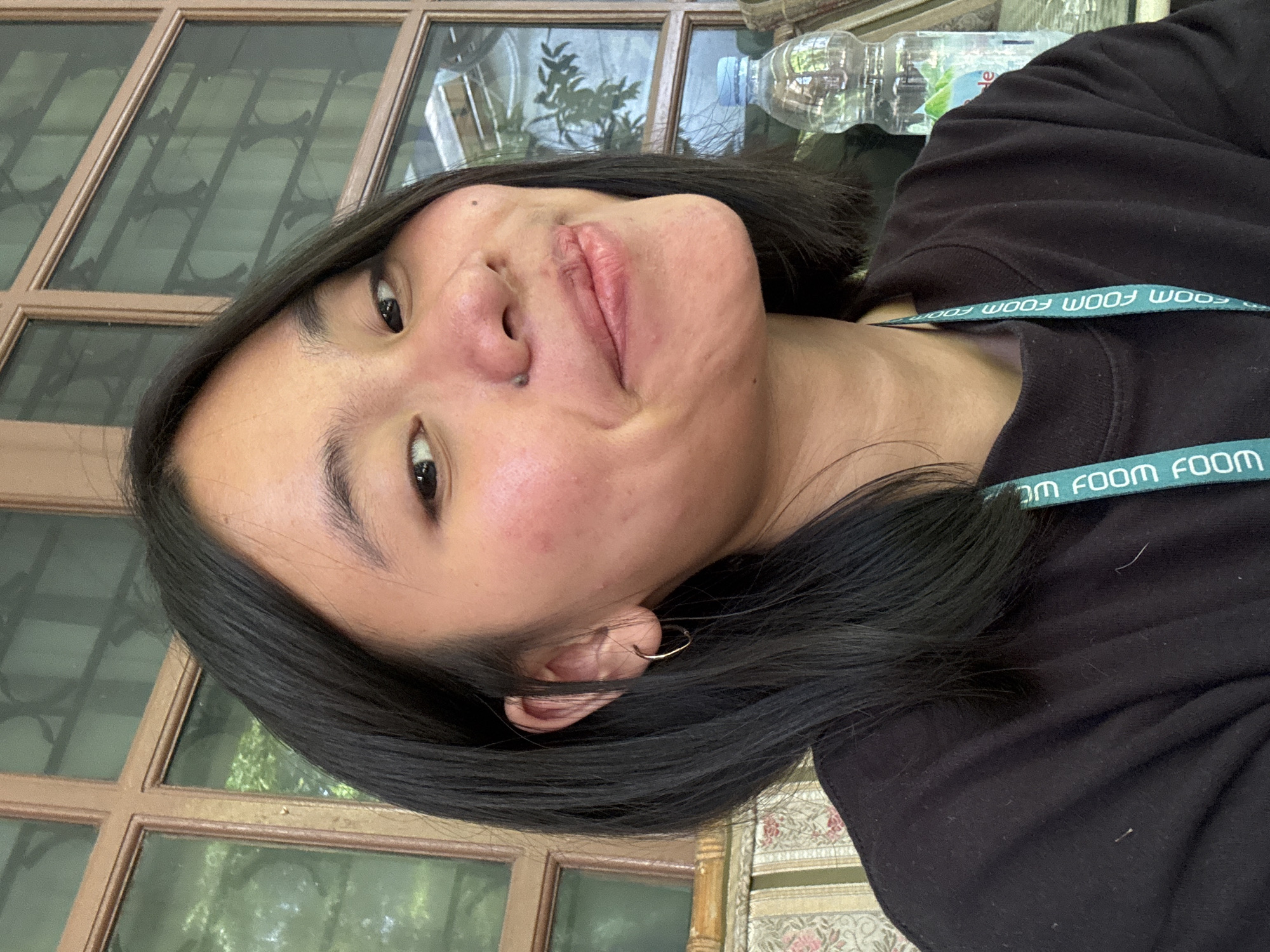 A young woman with dark, shoulder-length hair and a black t-shirt is pictured from the chest up. She is indoors, seated in front of a large window with wooden frames and tinted glass. The window overlooks greenery, suggesting an outdoor setting beyond the glass. To her right, a plastic water bottle sits on a surface. Her expression is a slight smile, and she is looking towards the camera. A teal lanyard with white text is visible around her neck. The background suggests a casual, possibly outdoor seating area with patterned upholstery on a bench. There is no indication of this being in South Jakarta, Indonesia.