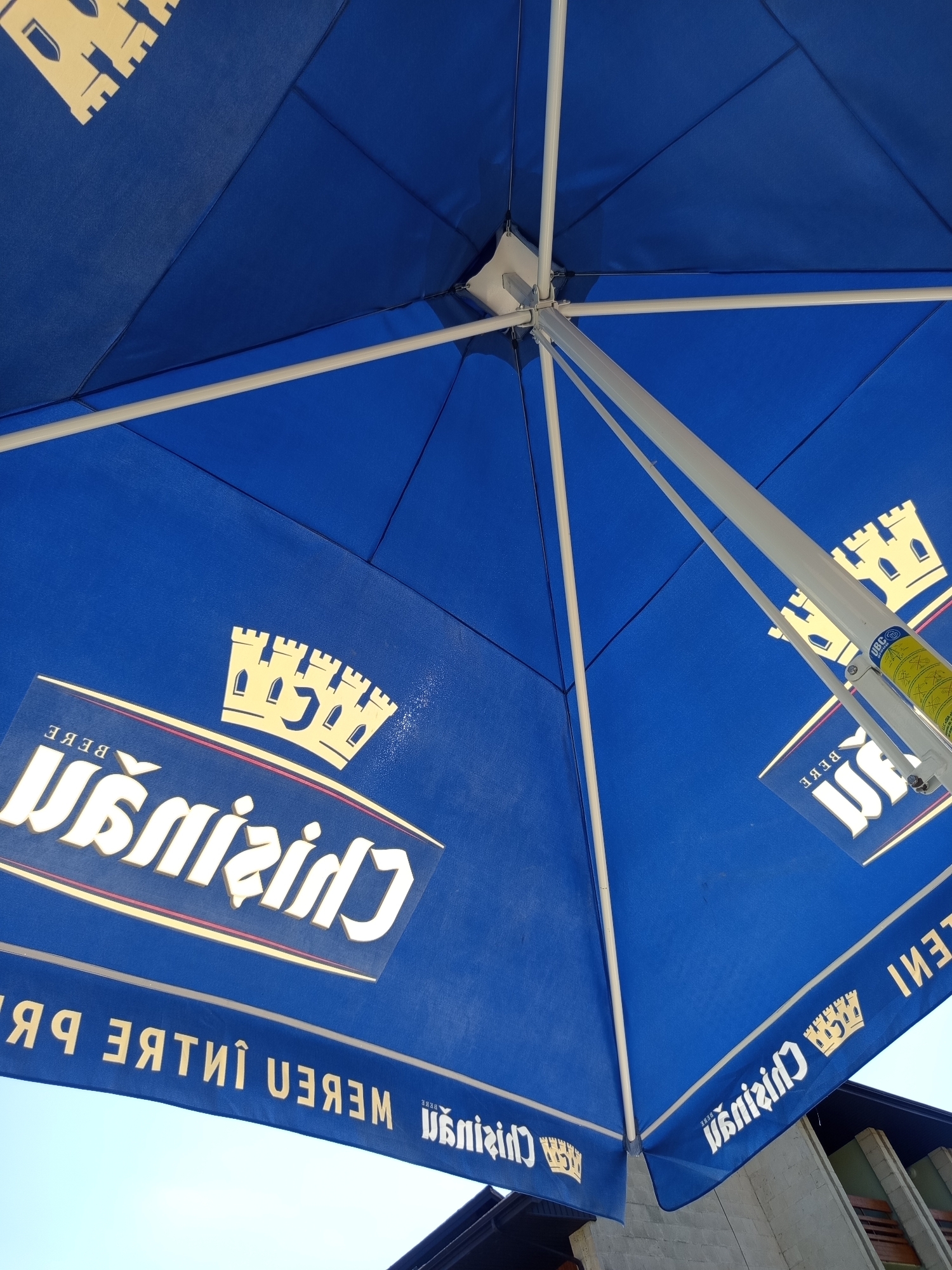 This image shows an underside view of a large, blue outdoor umbrella, likely a branded promotional item for "Crisan" beer. The umbrella is supported by a white metal frame and pole. Two identical logos are visible on the umbrella's fabric panels. Each logo features a crown symbol and the word "Crisan" in white lettering, with "BEBE" above it. There is a second, longer banner with text that appears to be the same brand name and "WEBER PR." The umbrella is positioned outdoors, with a bright sky visible in the background. The architecture in the background suggests a building or structure, possibly a cafe or restaurant, in Molovata Nouă, Moldova. The image is taken from a low angle, looking up at the umbrella.