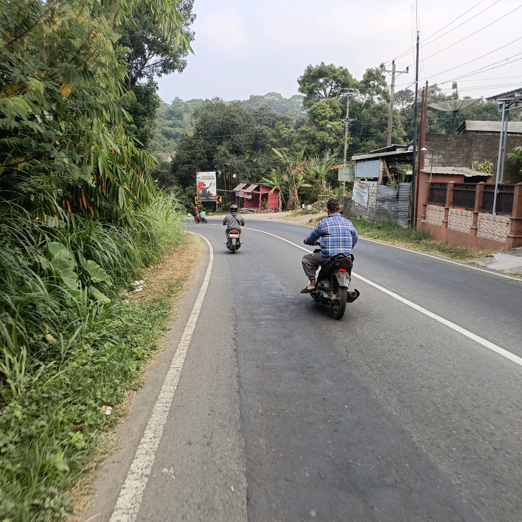 The image depicts a two-lane asphalt road in Sumogawe, Indonesia, curving through a lush, green environment. Tall trees and dense foliage line the left side of the road, with a white dashed line marking the edge of the pavement. On the right side, there are buildings and structures, including a brick wall with a decorative railing, and a satellite dish visible on a rooftop.
Two individuals are riding motorcycles on the road. The person in the foreground, on the right side of the image, is riding a black motorcycle and is wearing a blue and white checkered long-sleeved shirt and grey pants. They are facing away from the camera. Further ahead, on the left side of the road, another person is riding a motorcycle, also facing away from the camera, and is wearing a grey hooded sweatshirt and dark pants. A third, smaller motorcycle is visible in the distance, also moving away from the camera.
Along the roadside, there are some makeshift structures, including a small red building with a dark roof. A billboard advertising "REPUBLIK INDONESIA" with a prominent image of a man in traditional attire is also visible on the left side of the road. Electrical poles and wires are present throughout the scene, indicating the presence of infrastructure. The sky is overcast, suggesting a cloudy day. Some litter is scattered along the grassy verge on the left side of the road.
