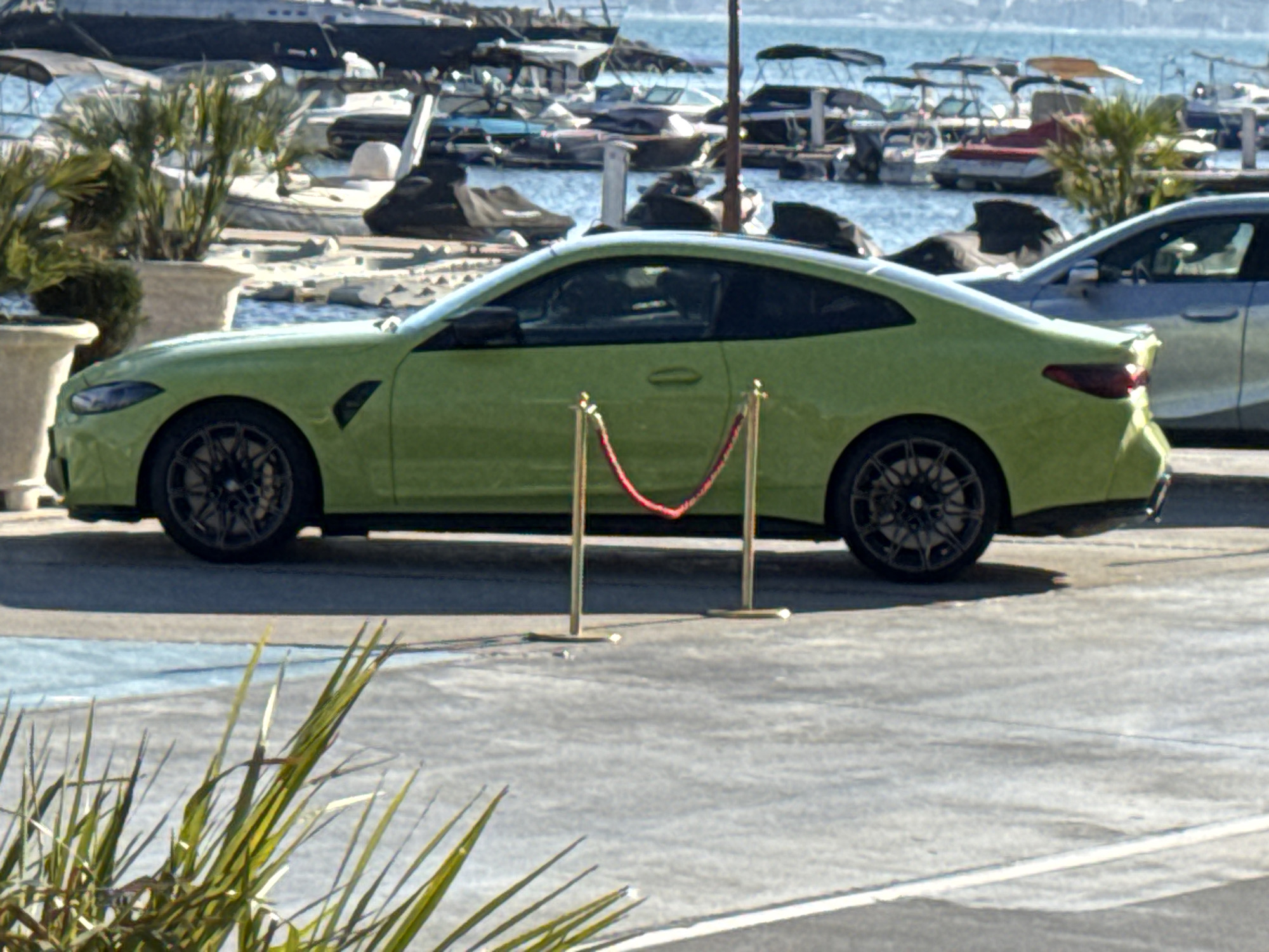 A lime green coupe is parked on a paved surface next to a body of water. The car is facing right, and its side profile is visible. It has black rims with a multi-spoke design. Behind the car, there is a line of boats docked in the water, and beyond that, a hazy coastline with hills. In the foreground, out-of-focus green foliage partially obscures the view. A stanchion with a red rope is positioned in front of the car, suggesting a barrier or an event setup. The sun appears to be shining, casting shadows, and the sky is a light blue. The scene looks like a sunny day at a harbor or marina. There is no visible text in the image.