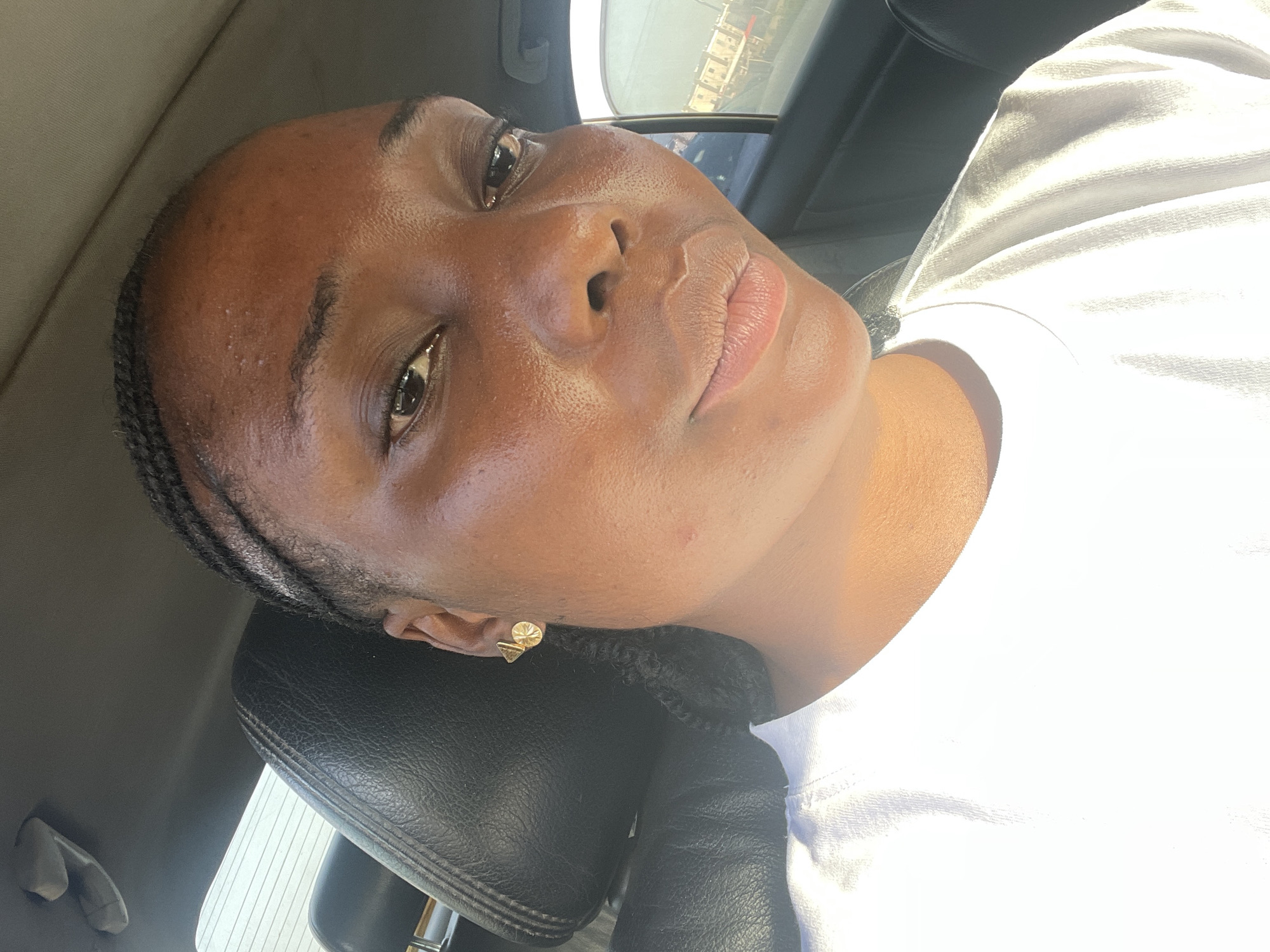 The image depicts a dark-skinned woman with braided hair and a small gold earring in a car. She wears a white shirt and rests her head on the black leather car seat headrest. The interior of the car is visible, with the window showing a blurry cityscape, possibly located in Gwarinpa, Nigeria.