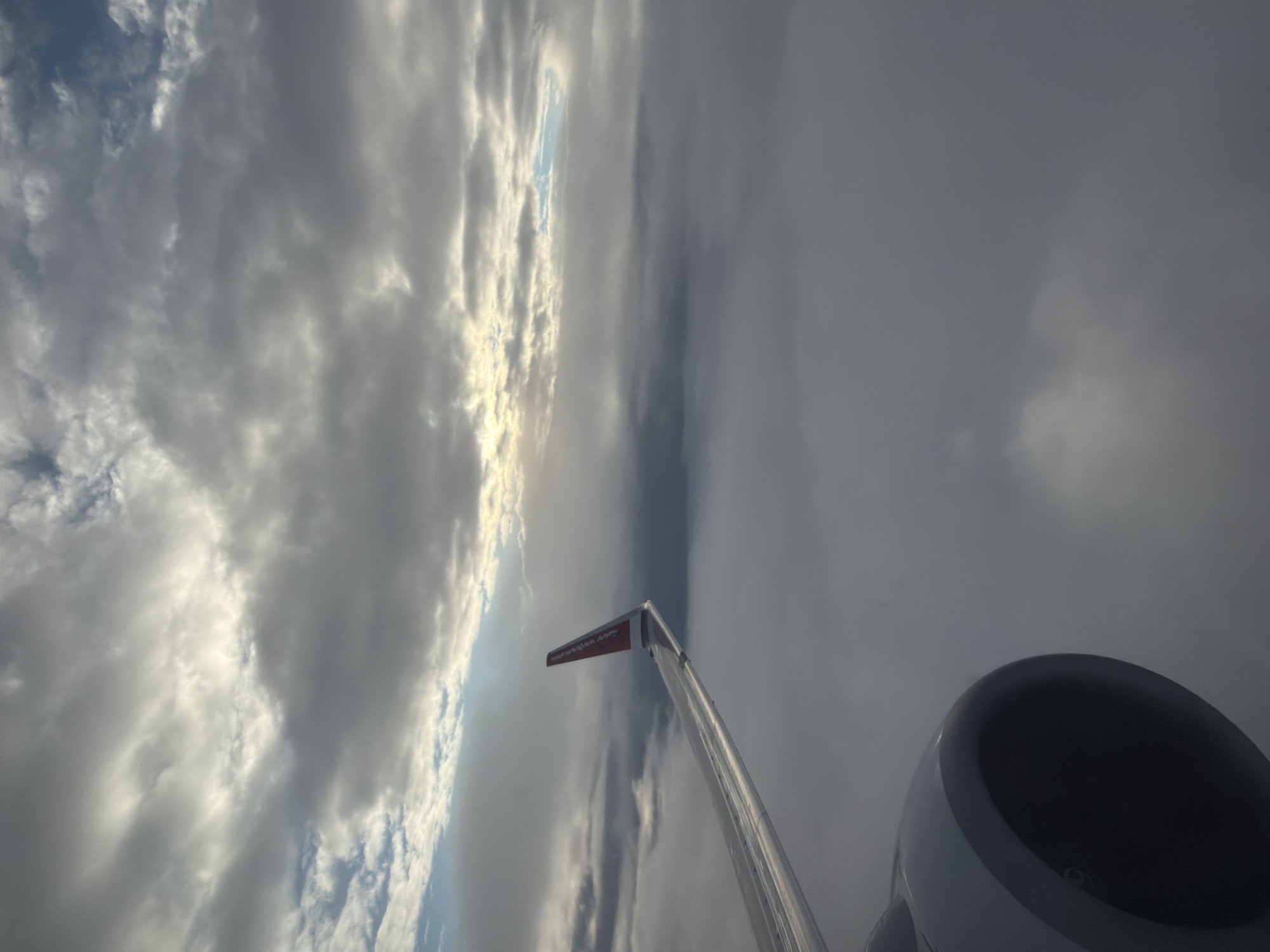 An aerial view from an airplane window shows a cloudscape with the wing and engine of the aircraft visible. The wing, which has a red tip with the Norwegian airline logo, extends from the left side of the frame. The engine, a dark cylindrical object, is partially visible in the lower left corner. The sky is filled with layers of gray and white clouds, with patches of brighter sky suggesting sunlight breaking through. The image does not provide any information about the specific location in Valle, Norway, or any people present.