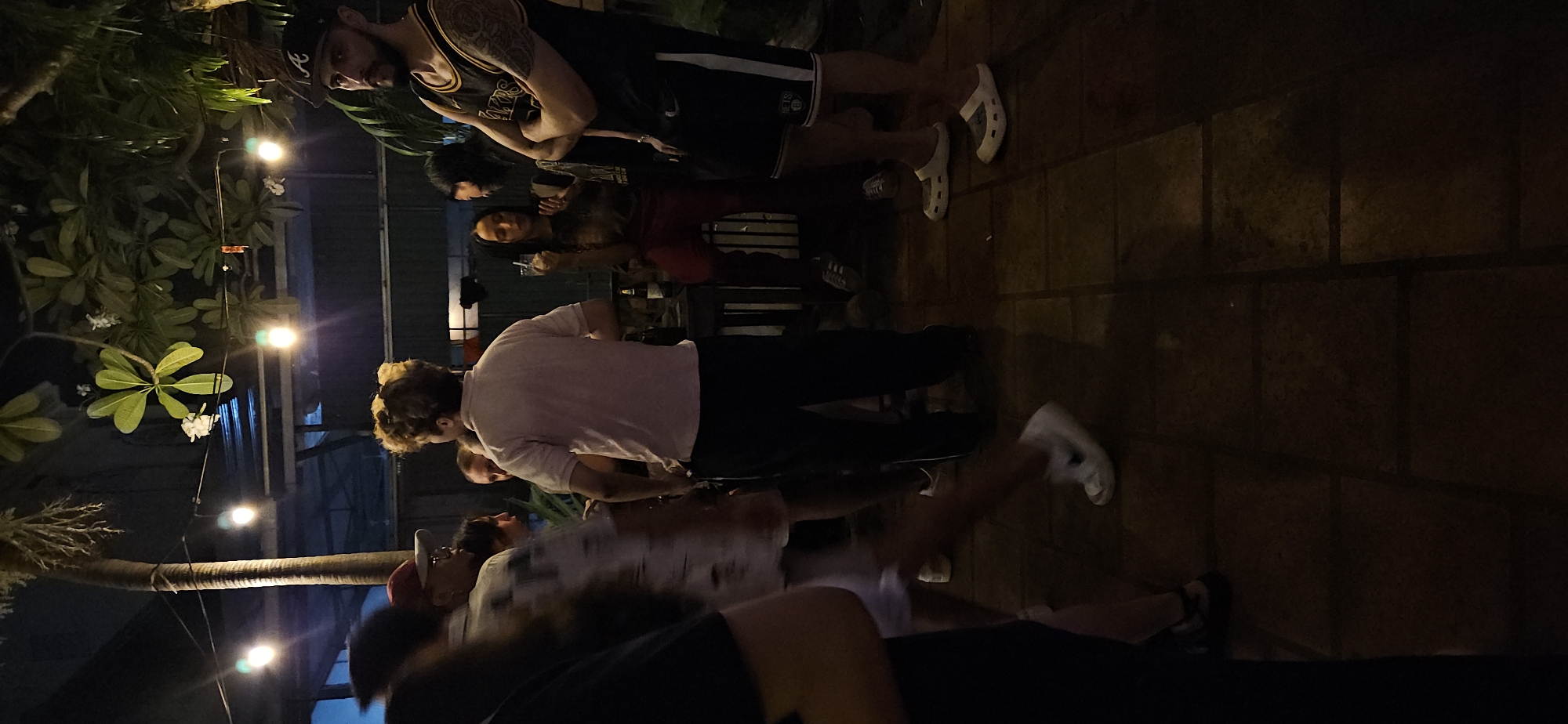 The image depicts an outdoor nighttime gathering, likely at a casual venue such as a bar or restaurant patio in Nha Trang Ward, Vietnam. Several individuals, mostly young adults, are standing and socializing under the dim, warm light of string bulbs.
In the foreground, a man with a dark beard and a black baseball cap, featuring a white 'A' logo, wears a black basketball-style tank top with text that appears to be "LAKERS" and black shorts with "NETS" printed on them. He stands with his arms crossed. Nearby, another man with curly blonde hair is seen from behind, wearing a light-colored t-shirt and dark pants. Further back, a woman with long dark hair, dressed in a dark top and reddish-brown pants, is partially visible, holding a drink. Other blurred figures suggest a larger group.
The environment features tropical plants, including frangipani with white flowers, and a prominent palm tree trunk on the left side of the frame. The ground is paved with dark, square-patterned tiles. Artificial string lights crisscross the scene, providing the primary illumination and indicating it is nighttime. The atmosphere appears relaxed and social. One person is wearing distinct white Crocs. Beyond the logos on clothing, no other discernible text is visible.