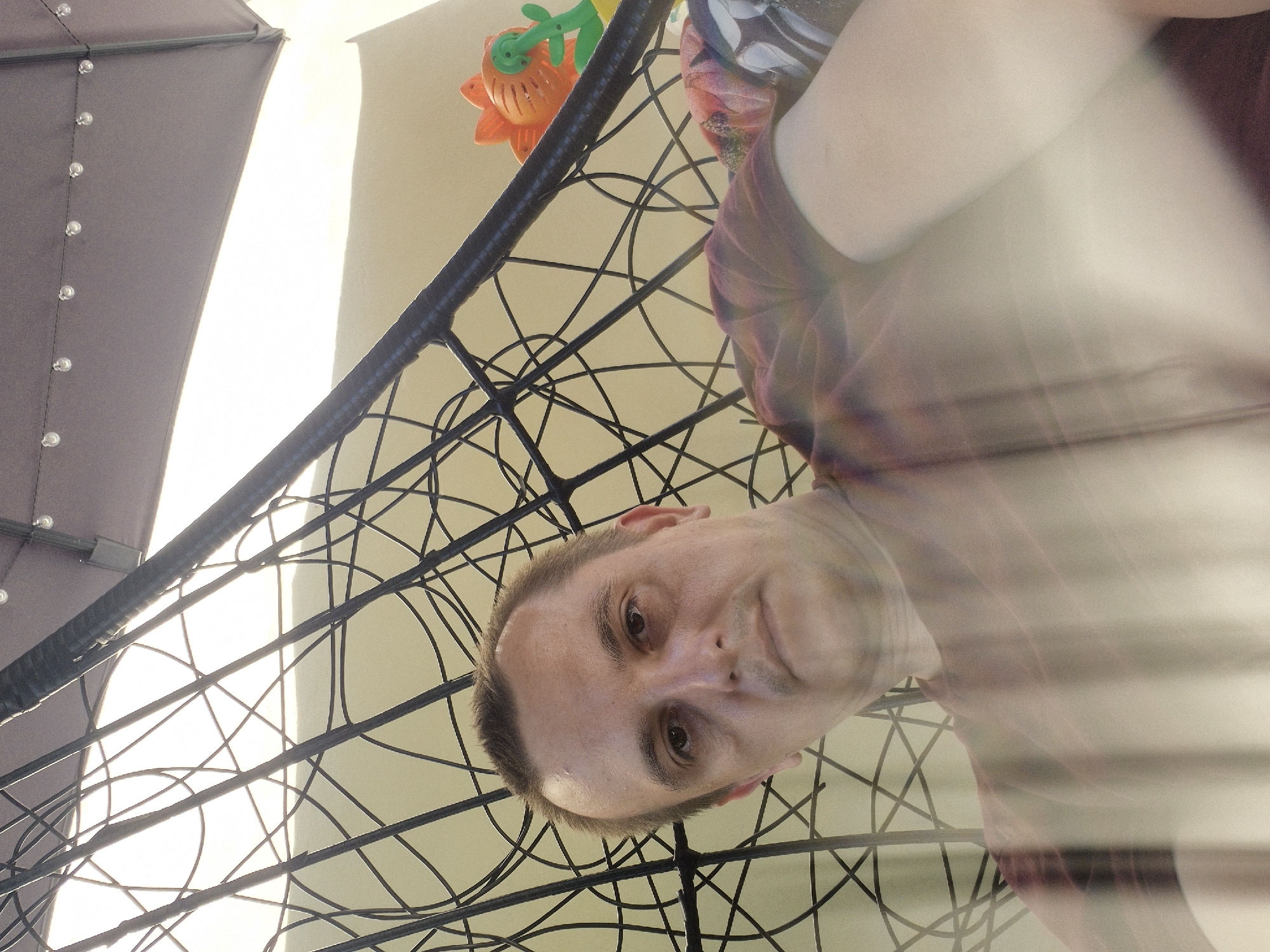 This is a close-up, low-angle selfie of a man's face and upper body, taken outdoors. He is looking directly at the camera with a neutral expression. His short brown hair is slightly messy. He is wearing a maroon t-shirt.
The man is positioned in front of a black, woven structure, possibly a chair or a hammock, which forms a complex, intersecting pattern in the foreground. Behind him, a large, dark object with visible silver studs, likely an umbrella or canopy, is partially visible on the left. The background is bright, suggesting daylight, with a pale, possibly wall-like surface. A colorful toy, shaped like an orange flower with green accents, is also visible in the upper part of the image, hanging from the woven structure. The overall lighting is bright and appears to be natural sunlight, indicating it is daytime. The scene suggests a casual outdoor setting, possibly a patio or backyard in Švenčionys, Lithuania. There is no visible text in the image. The motion blur in some parts of the image indicates movement during the photograph.