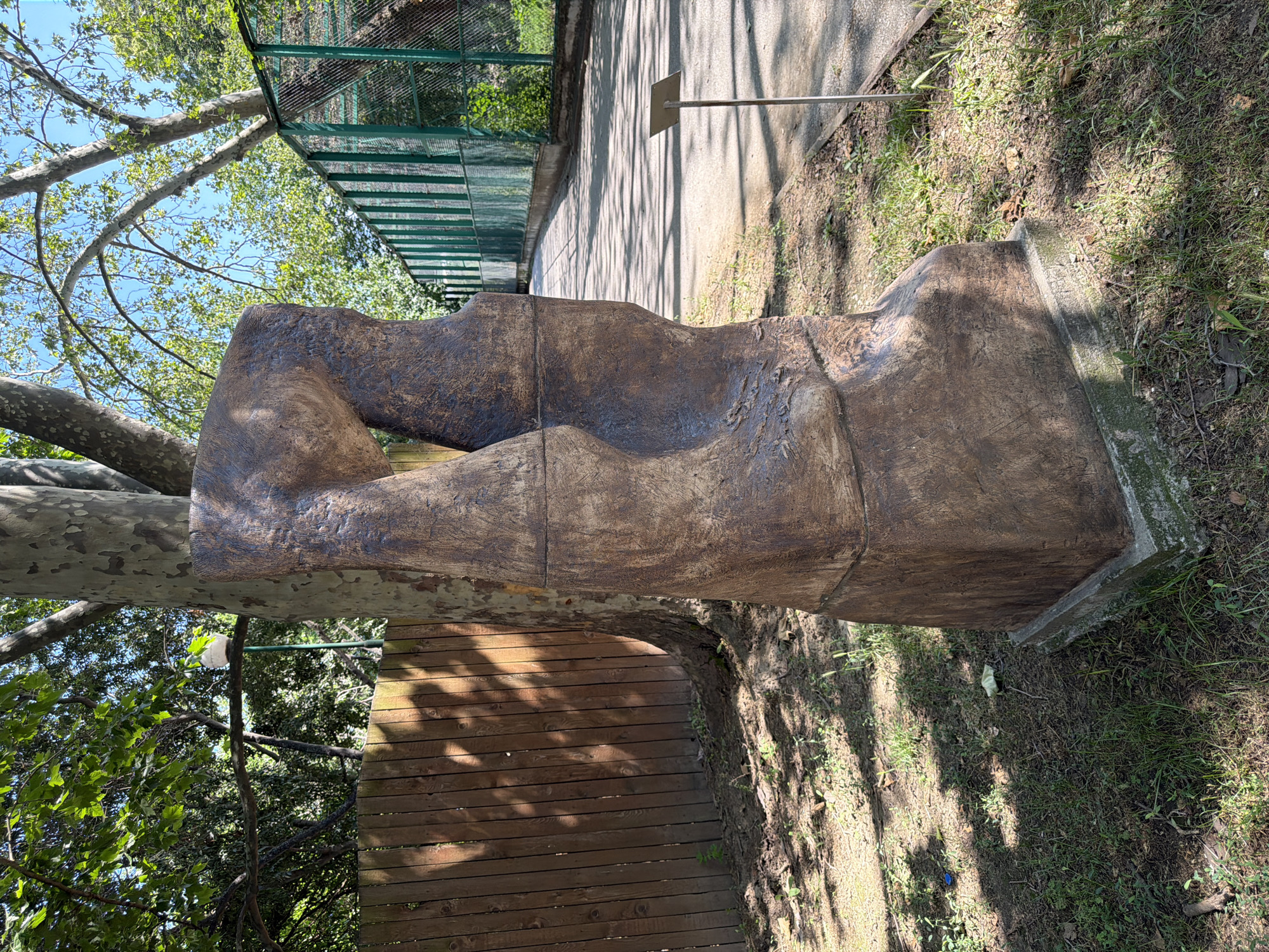 The image shows a large, abstract sculpture made of a rough, textured material resembling weathered wood or stone. The sculpture has a dark brown color and is roughly rectangular in shape, with curved indentations that give it an organic, almost anatomical appearance. It is placed outdoors on a grassy area with a concrete path running alongside it, casting dappled shadows from the surrounding trees.
In the background, a green metal fence encloses an area with more trees and foliage. A silver metal pole with a small, square sign is planted in the ground near the sculpture. The overall setting appears to be a park or an outdoor sculpture garden, possibly within a zoo or animal enclosure, given the presence of the fencing.
The sun is shining, creating distinct shadows, suggesting it is daytime. The presence of lush green leaves on the trees indicates a warm season, likely spring or summer. The sculpture itself is the primary subject, and its abstract form invites interpretation. No people are visible in the image. The scene is peaceful and natural, with the sculpture integrated into the environment.