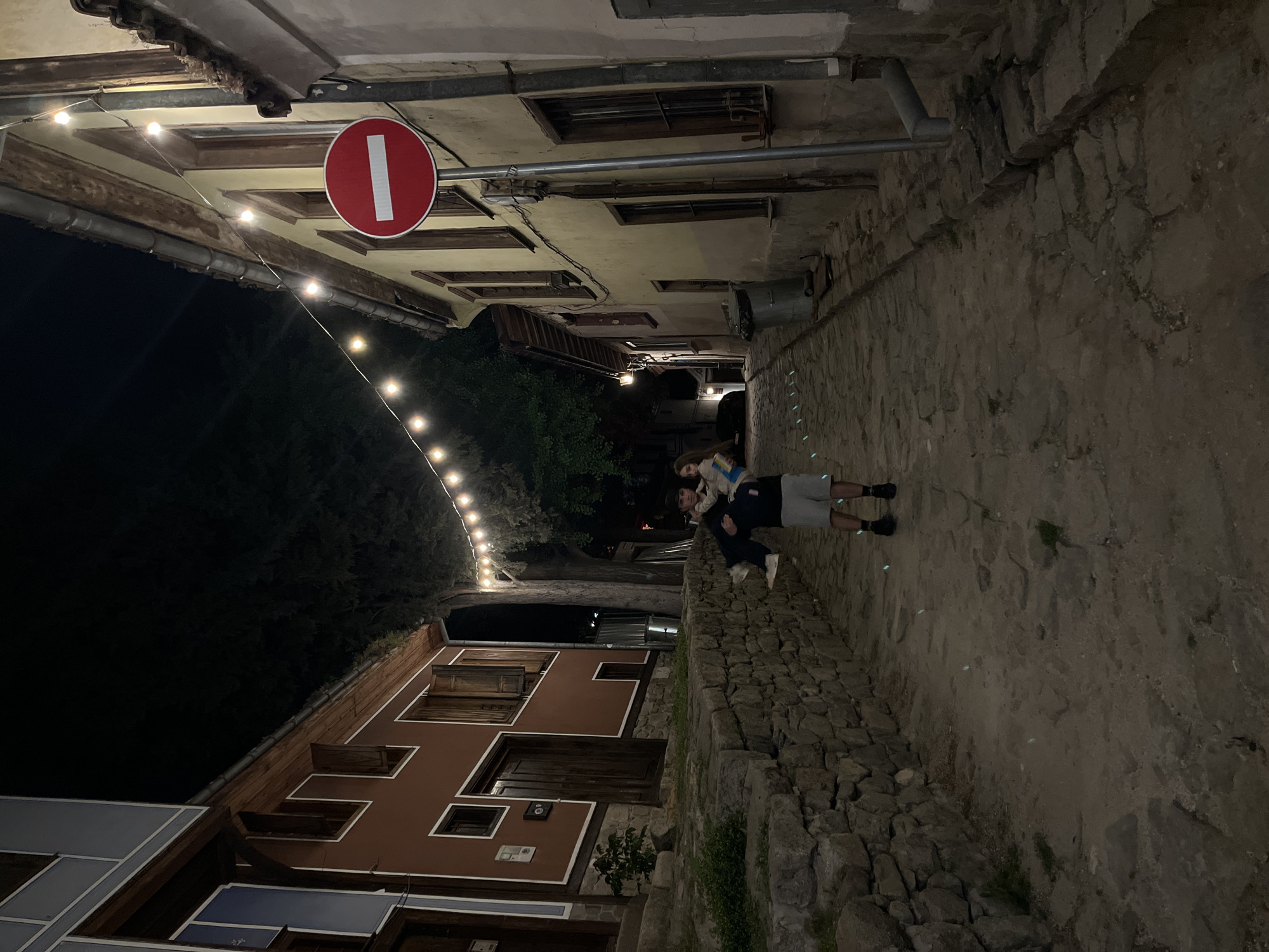 A nighttime scene in Plovdiv, Bulgaria, shows a narrow cobblestone street lit by string lights, with old buildings lining both sides. A no-entry sign is visible overhead. Two people stand in the street. One man is crouched down and the other is sitting on his shoulders.