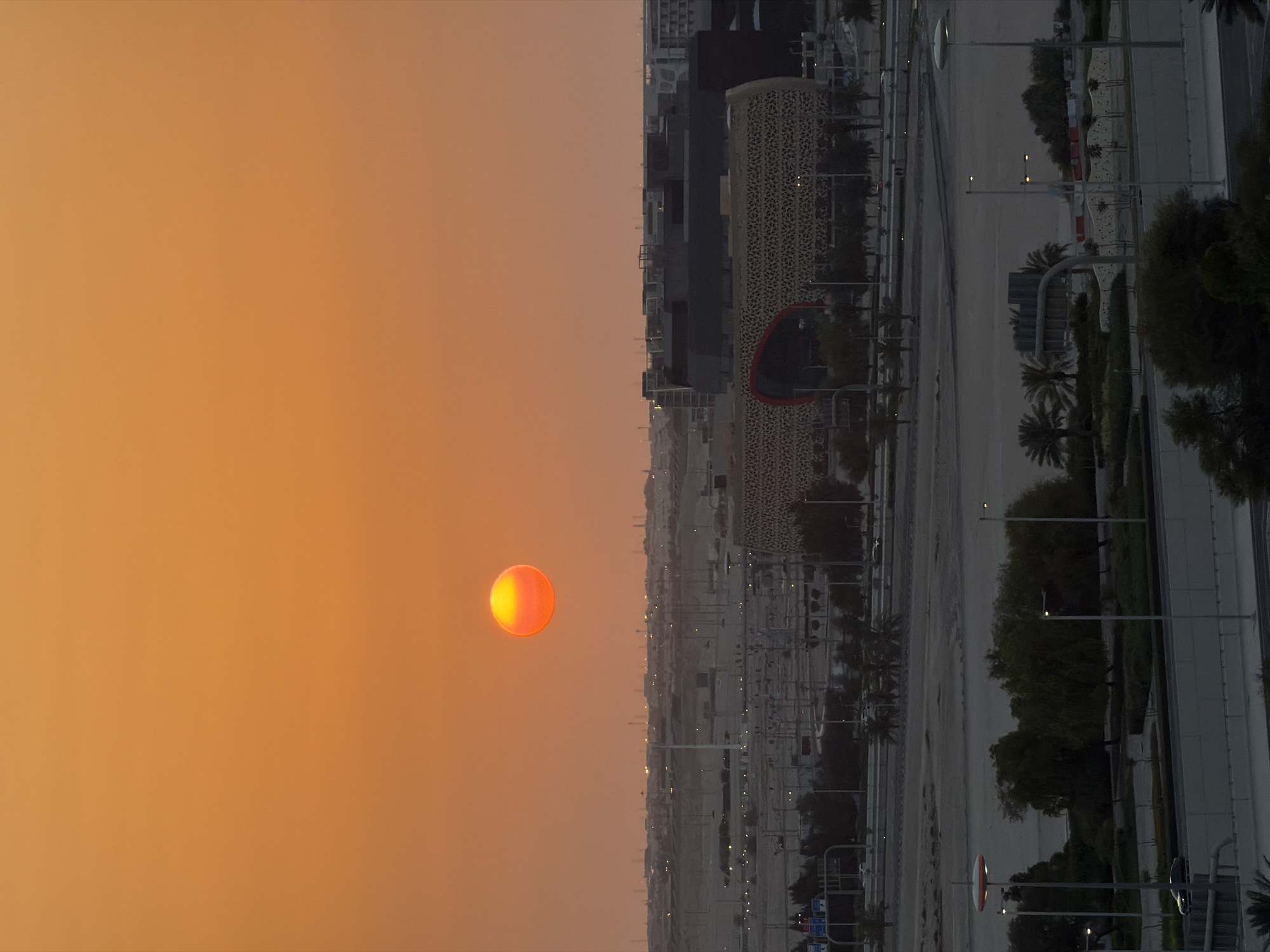 An aerial shot captures the sunset over an urban landscape. The sun, a vibrant orange sphere, is positioned in the upper left quadrant of the frame, casting a warm glow across the sky. Below, a city stretches towards the horizon, characterized by low-rise buildings and structures with unique, patterned facades. Roads and infrastructure are visible, suggesting a developed area. The foreground is dominated by greenery, including trees and bushes, with streetlights dotting the scene. The overall impression is one of a developing urban environment at dusk.