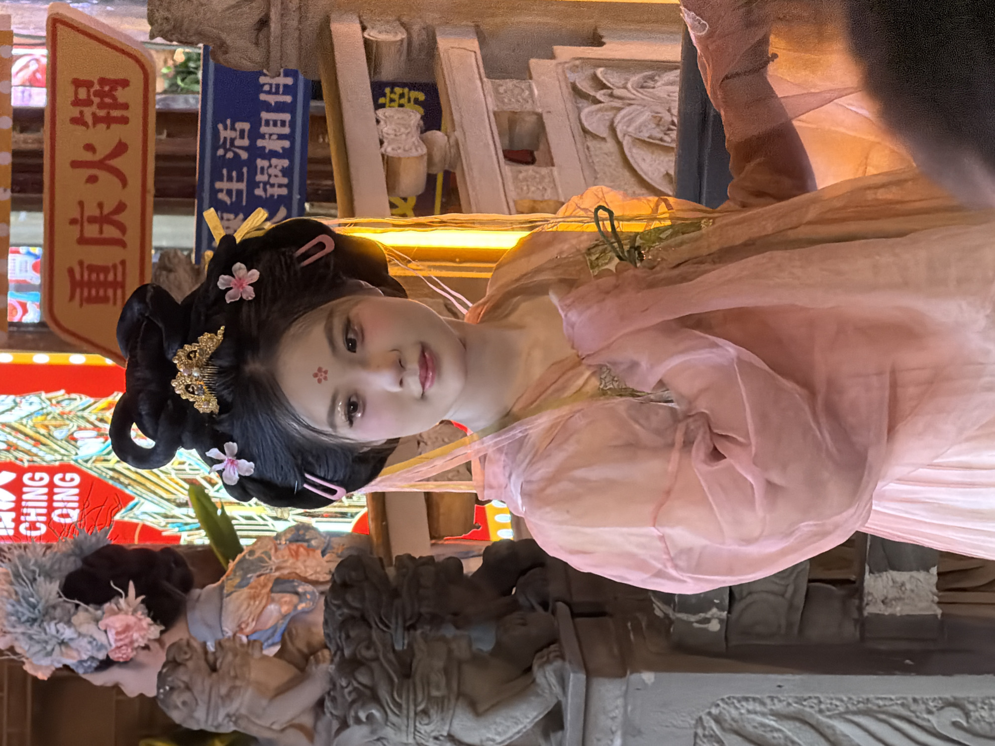 A young East Asian woman, adorned in traditional Chinese attire and makeup, is posed in front of a backdrop that suggests a commercial establishment. She wears a black, ornate updo with decorative hairpins and a pink hair accessory. Her makeup features rosy cheeks and subtle eye embellishments. Her garment is a flowing, sheer pink robe over a lighter pink dress, with decorative elements around the neckline.
Behind her, a sign with Chinese characters, "重庆火锅" (Chóngqìng huǒguō - Chongqing Hot Pot), is visible, indicating the likely location of a restaurant. Another blue sign with Chinese text is also present. To the left of the woman, a stone lion statue is partially visible, a common decorative element in Chinese culture. The overall scene appears to be set within a street or market area, possibly at night, given the lighting and the illuminated signs. The background elements suggest the setting is within the Yuzhong District of Chongqing, China, a well-known area for its bustling street life and vibrant restaurant scene.