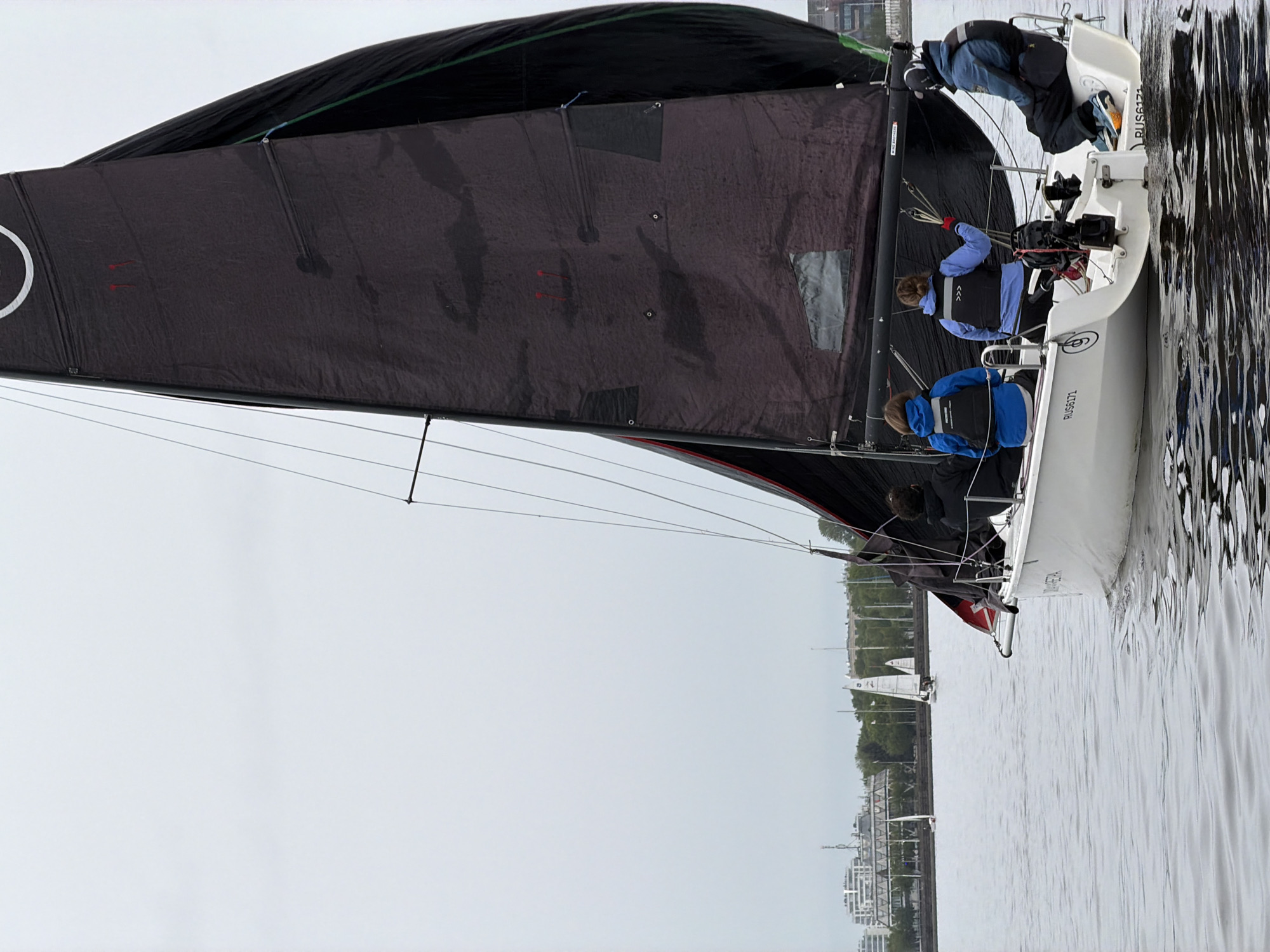 A sailboat with a black sail is underway on a body of water, with the city of Saint Petersburg, Russia, visible in the background. The sailboat has at least three people on board, wearing life vests. Two people are closer to the stern of the boat, with one appearing to be steering or handling lines. Buildings and trees line the shoreline in the distance. The sky is overcast and the water is relatively calm. The boat's hull displays identification markings "RUS6171".