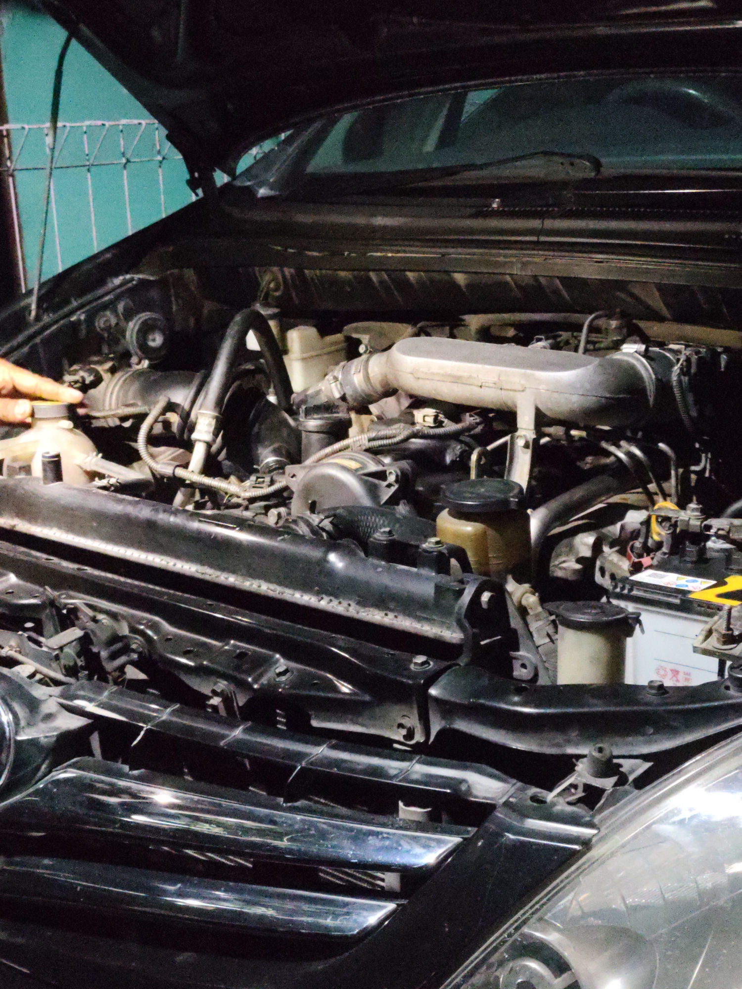 The image shows the engine bay of a black car, likely in an outdoor or semi-outdoor setting given the lighting conditions and the glimpse of a fence in the background. A person's hand is visible on the left side of the frame, touching the cap of a translucent fluid reservoir. The engine components, including various hoses, pipes, a large air intake, and the battery, are clearly visible. The hood of the car is open, revealing the engine. The overall impression is that of routine car maintenance or inspection. Based on the provided context, this scene is situated in Bulakrejo, Indonesia.