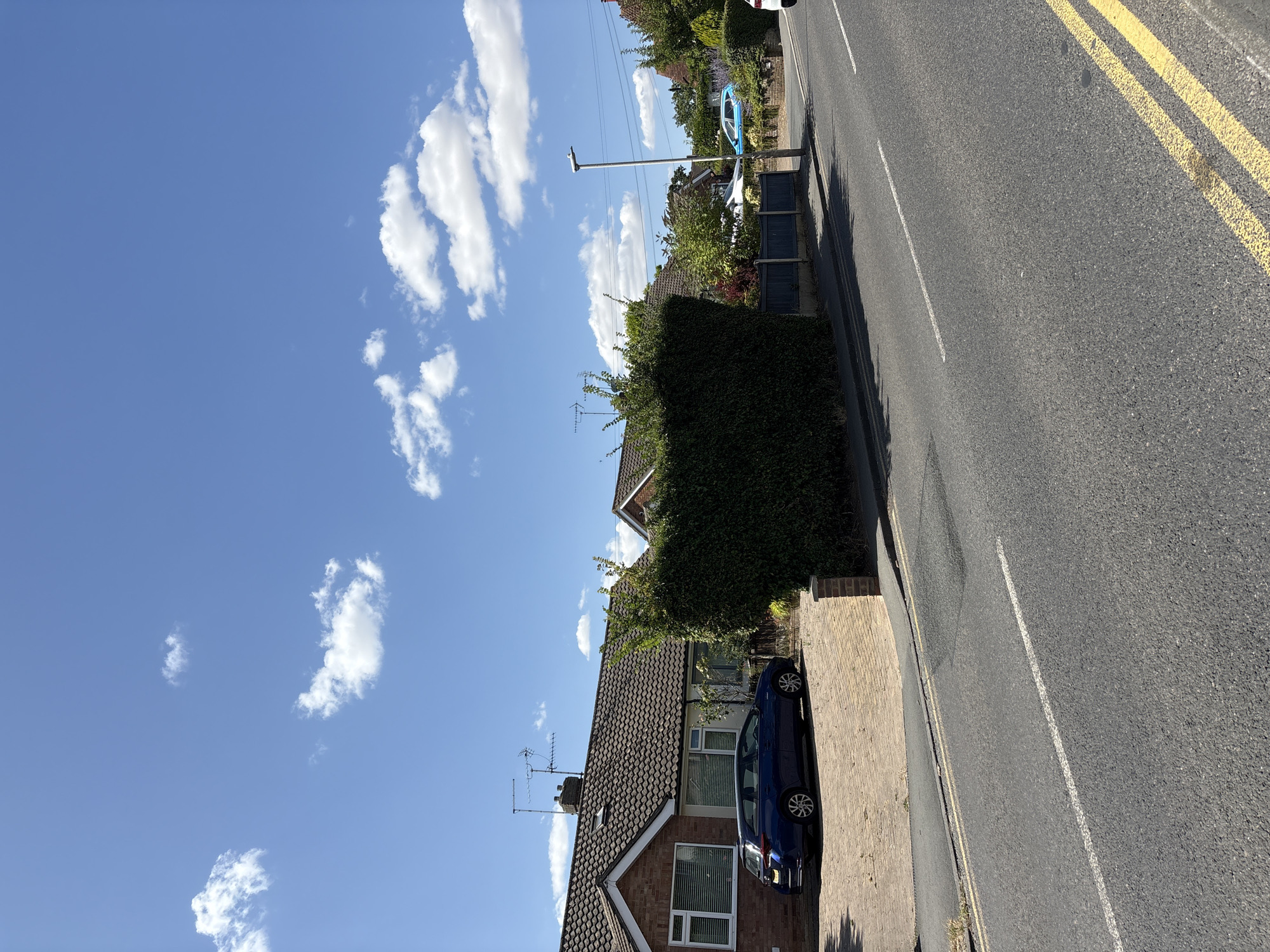 The image depicts a suburban street scene under a bright blue sky with scattered white clouds. In the foreground, a paved road runs diagonally from bottom left to top right, marked with a white line and double yellow lines on the right side. On the left side of the road, a driveway paved with light-colored bricks leads to a single-story brick house with a tiled roof. A dark blue station wagon is parked on the driveway. A tall, dense green hedge runs along the boundary between the driveway and the pavement. Beyond the hedge, a dark blue fence separates the property from the street. Further down the street, more houses and trees are visible. A grey lamppost stands on the pavement. In the distance, a white car and a light blue car are parked on the side of the road. The overall impression is a sunny day in a residential area.