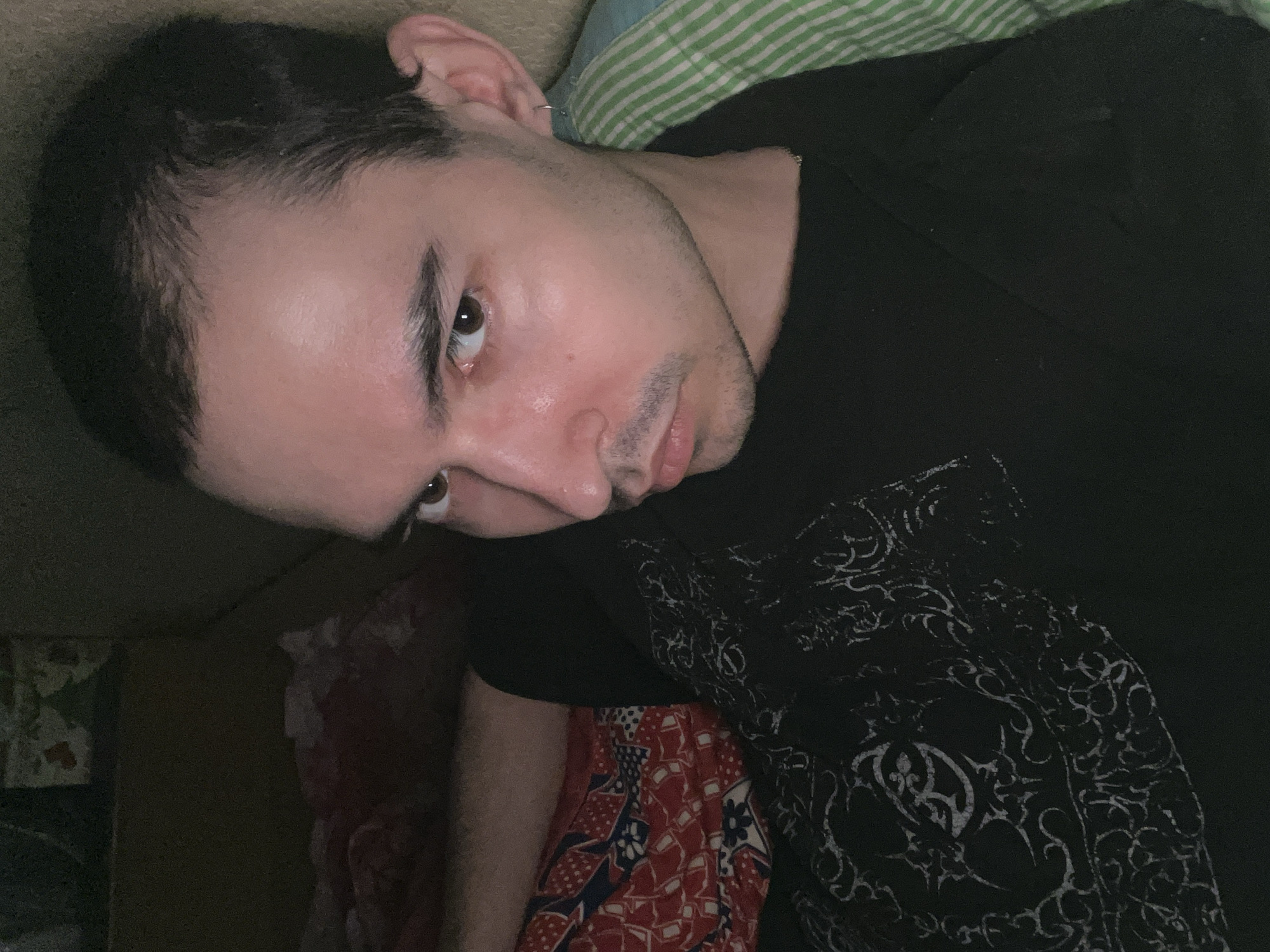 A medium shot shows a young adult male from the chest up, lying down, looking to the viewer's right. The man has short dark hair, dark eyebrows, and a light complexion with some shine on his forehead and cheeks. He is wearing a black t-shirt with a white, intricate pattern on the left side. His left ear has a small silver hoop earring. A green and white striped fabric is visible behind his head on the right. The background is dark and indistinct, suggesting an indoor setting, possibly a bedroom. The image is taken from a slightly high angle.
