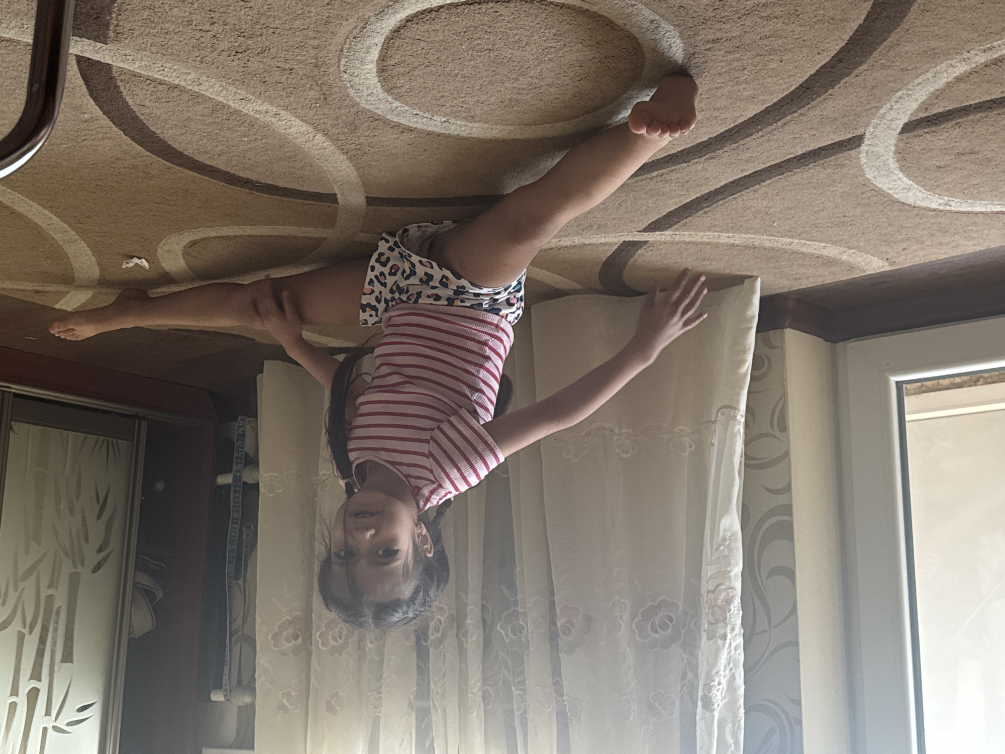 An indoor scene in a residential dwelling, possibly a living room or bedroom, located in the city of Unknown, Unknown. A young girl, approximately 5-8 years old, with dark hair styled in braids, is performing a gymnastic split on a light brown carpet. She wears a pink and white striped short-sleeved shirt and white shorts with a multi-colored animal print pattern. Her legs are fully extended to her sides, and her torso is upright, turned slightly towards the viewer. Her right hand is flat on the carpet, while her left hand is on her left thigh. She looks directly at the camera with a neutral expression.
The carpet features a geometric pattern of large, intertwined dark brown and off-white curved lines. To the left, a white-framed window or glass door is visible, admitting bright natural light. Light-colored sheer curtains with embroidered floral patterns are drawn across the background, partially obscuring a light-colored wall with a subtle patterned wallpaper. To the far right, a dark brown wooden cabinet or wardrobe with a frosted glass panel featuring a bamboo motif is present. A white pillow is partially visible near the girl's right side, and a crumpled piece of white paper rests on the carpet near her right leg. A curved metallic object is partially visible in the bottom right corner of the frame.
