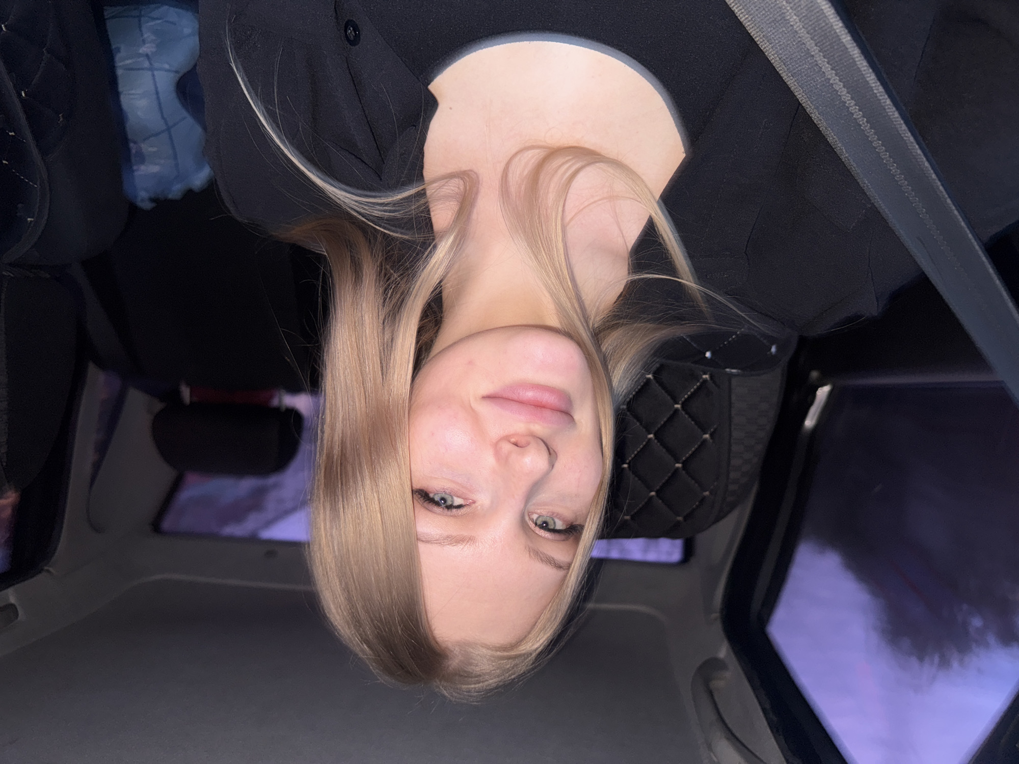 A selfie of a fair-skinned blonde woman with light eyes taken inside a car. She is wearing a black top and a seatbelt. The interior of the car is visible, including the dashboard, window, and a quilted seat cover. The location is unknown and cannot be determined from the image.