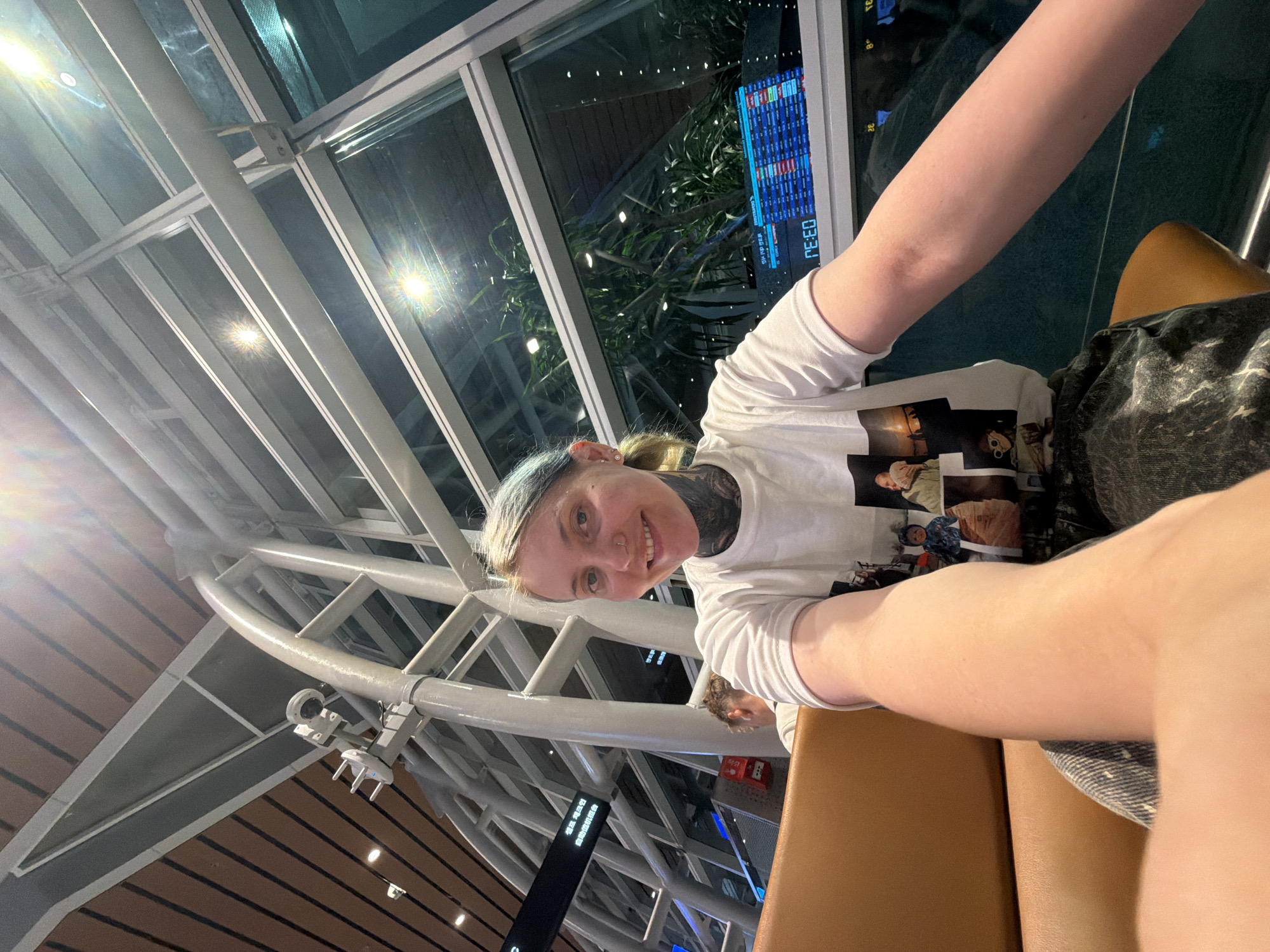 A selfie-style image captures a young person smiling in what appears to be a modern airport terminal in Đà Nẵng, Vietnam, at night. The subject, likely female, has light-colored hair with blue/green streaks, a nose piercing, and extensive dark tattoos visible on her neck and collarbone. She is wearing a white long-sleeved t-shirt featuring multiple photographic prints of people, and dark patterned shorts or a skirt. Her right arm is extended, holding the camera to take the photo.
The background reveals the distinctive architecture of a large public space, with a high ceiling composed of intricate silver metal beams and expansive glass panels. Through the windows, the outdoor area is dark, but reflections of bright internal lights are prominent. A large digital display screen, likely showing flight information, is visible through the glass. Partial text on this screen includes "03:30" and "03:32," indicating times, along with other alphanumeric characters related to flights. Lush green foliage is also visible through the window, suggesting landscaping outside.
Further details in the environment include a security camera or similar device mounted on one of the ceiling beams. Another dark, vertical informational sign with white text is present in the deeper background, though the text is unreadable. The subject's expression is one of pleasantness, with a clear smile directed at the camera. The setting strongly suggests an airport lounge or concourse during evening hours.