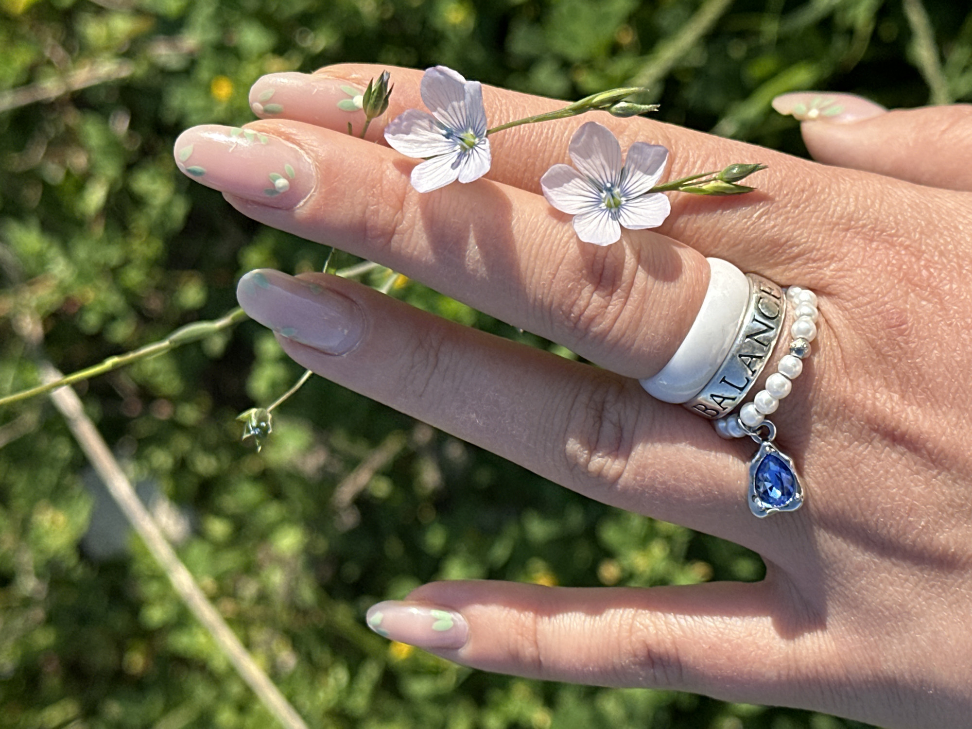 A close-up, outdoor photograph captures a human hand adorned with jewelry and delicate flowers, set against a blurred green background, presumably in Šušanj, Montenegro. The hand, with light-toned skin, displays manicured oval nails featuring a translucent nude polish with light-green heart or leaf-like designs. On the ring finger, stacked rings include a wide white band, a silver-toned band engraved with the word "BALANCE," and a beaded ring composed of small white pearls. A small silver-toned charm with a teardrop-shaped blue gemstone dangles from the pearl ring. Two light-lavender or pale-blue five-petaled flowers, resembling flax blossoms, are positioned against the index and middle fingers, their slender green stems bearing additional small green buds. The background is composed of out-of-focus green foliage, with distant hints of yellow flora.