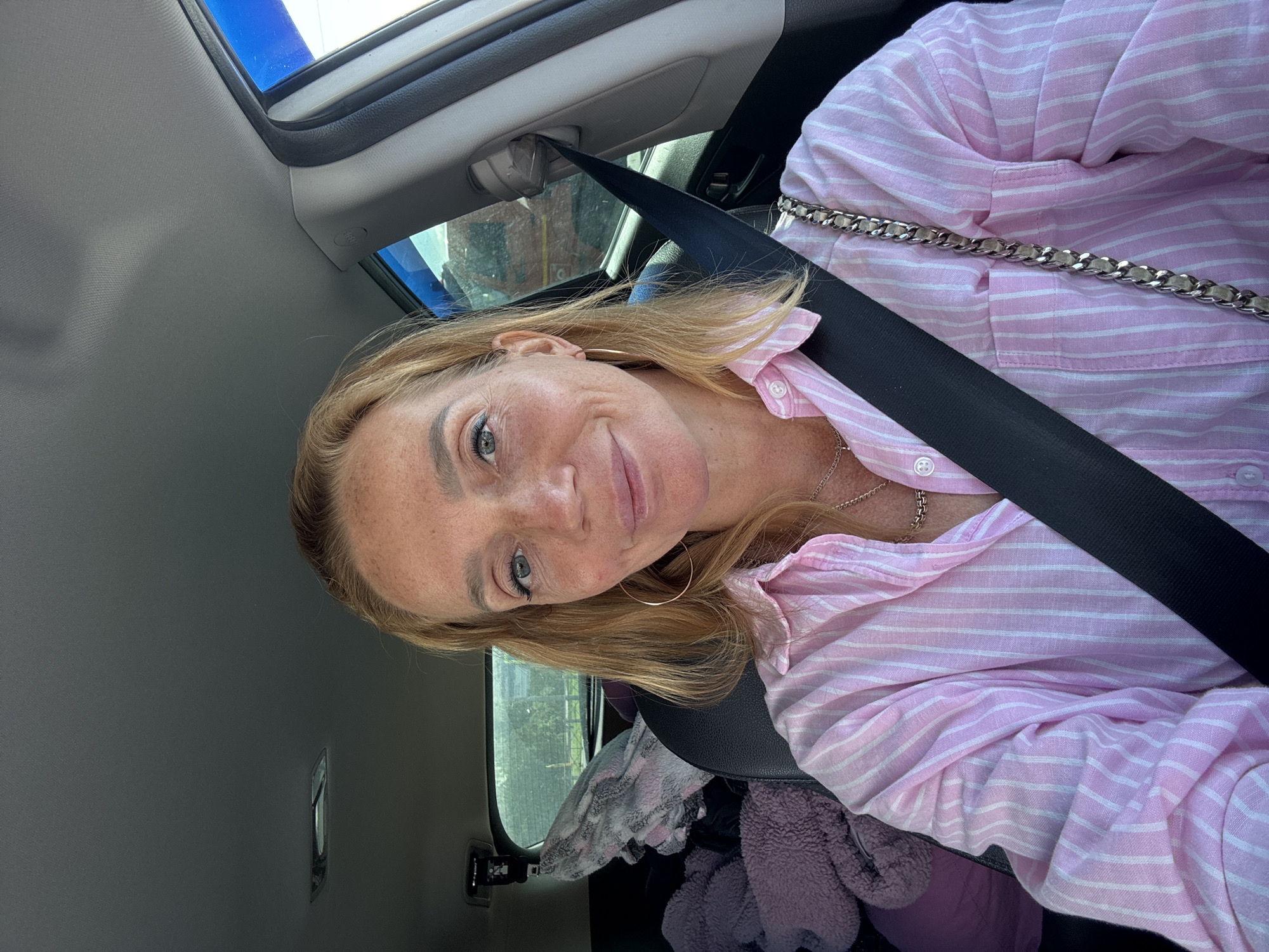 A woman with blonde hair and freckles is in the driver's seat of a car, wearing a pink and white striped shirt and a seatbelt. She is smiling and looking at the camera. The car's interior is visible, including the dashboard, ceiling, and back seat with stuffed animals and blankets. The background shows trees and a building through the car window. The image appears to be a selfie taken during the day.