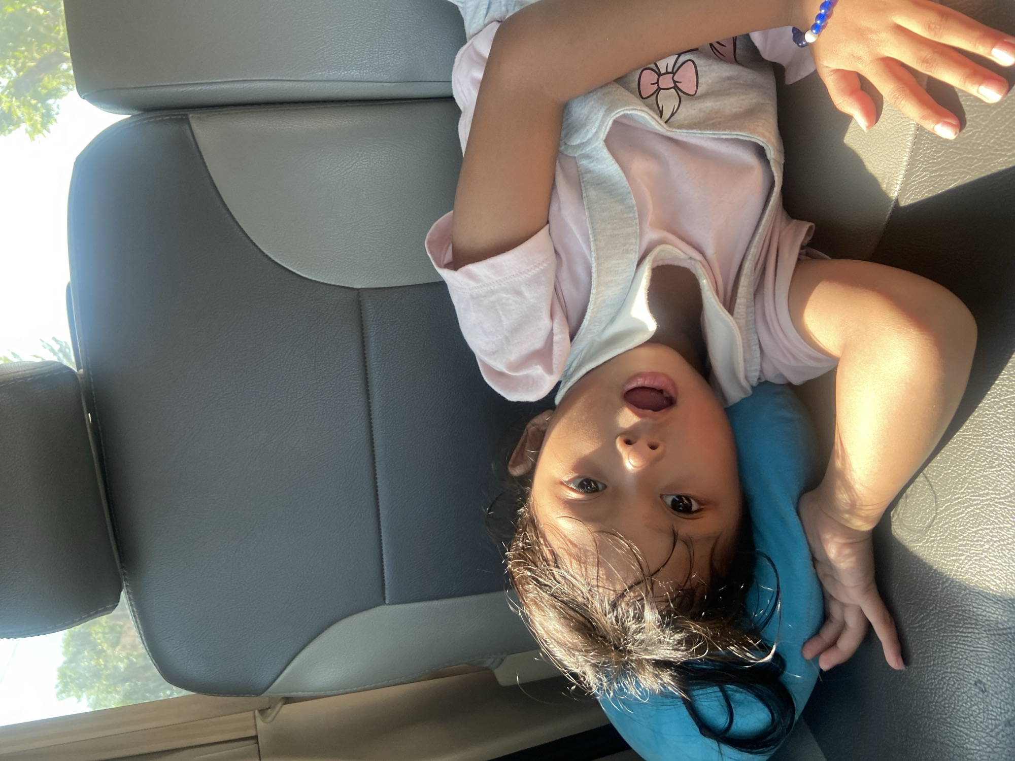A young child with dark hair lies on a blue pillow in the back seat of a vehicle. The child is wearing a pink t-shirt and overalls with a bow design. Sunlight streams in from the right side of the image, casting shadows on the child and the gray and tan leather seats. The background shows trees and sky through the vehicle window. The location is Ngrame, Indonesia.
