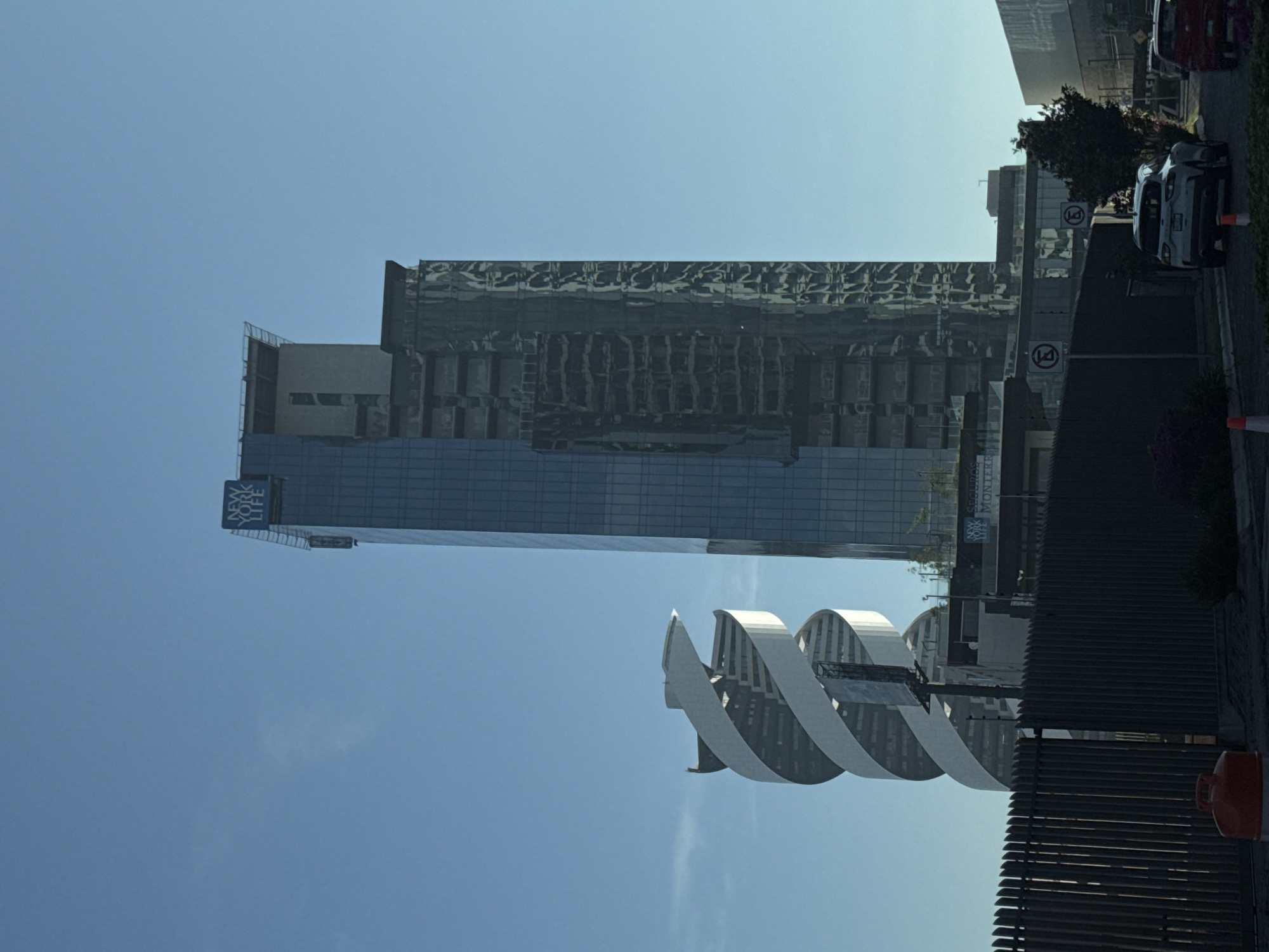 The image depicts a street scene in San Bernardino Tlaxcalancingo, Mexico, featuring modern architecture. Prominently visible are several high-rise buildings. One building features a reflective facade, while another displays the "New York Life Seguros Monterrey" logo. A swirling, white, architectural structure is in the foreground. Along the right side of the frame, there is a fence, shrubbery, traffic cones, and parked cars. Traffic signs are present. The sky is a clear blue. The scene suggests a contemporary urban environment.