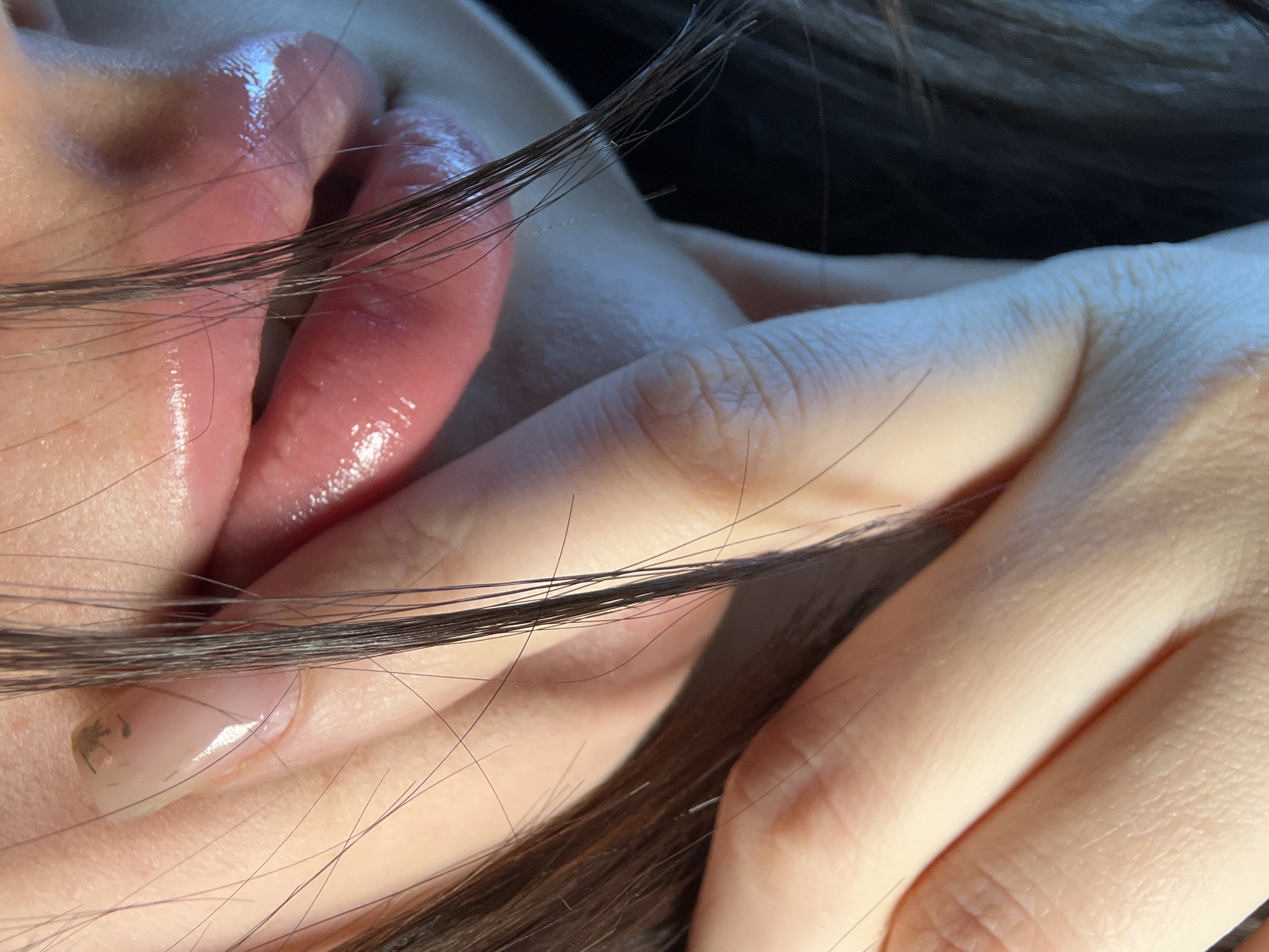 A close-up image shows a person's lips, fingers, and strands of dark hair. The lips appear glossy and are slightly parted. A hand is positioned with fingers curled, partially obscuring the lower portion of the face. The fingernail on the index finger has a pale pink hue with a small, dark imperfection near the tip. The background is dark and out of focus. There is no evidence in the image to indicate the location is Tyumen, Russia.