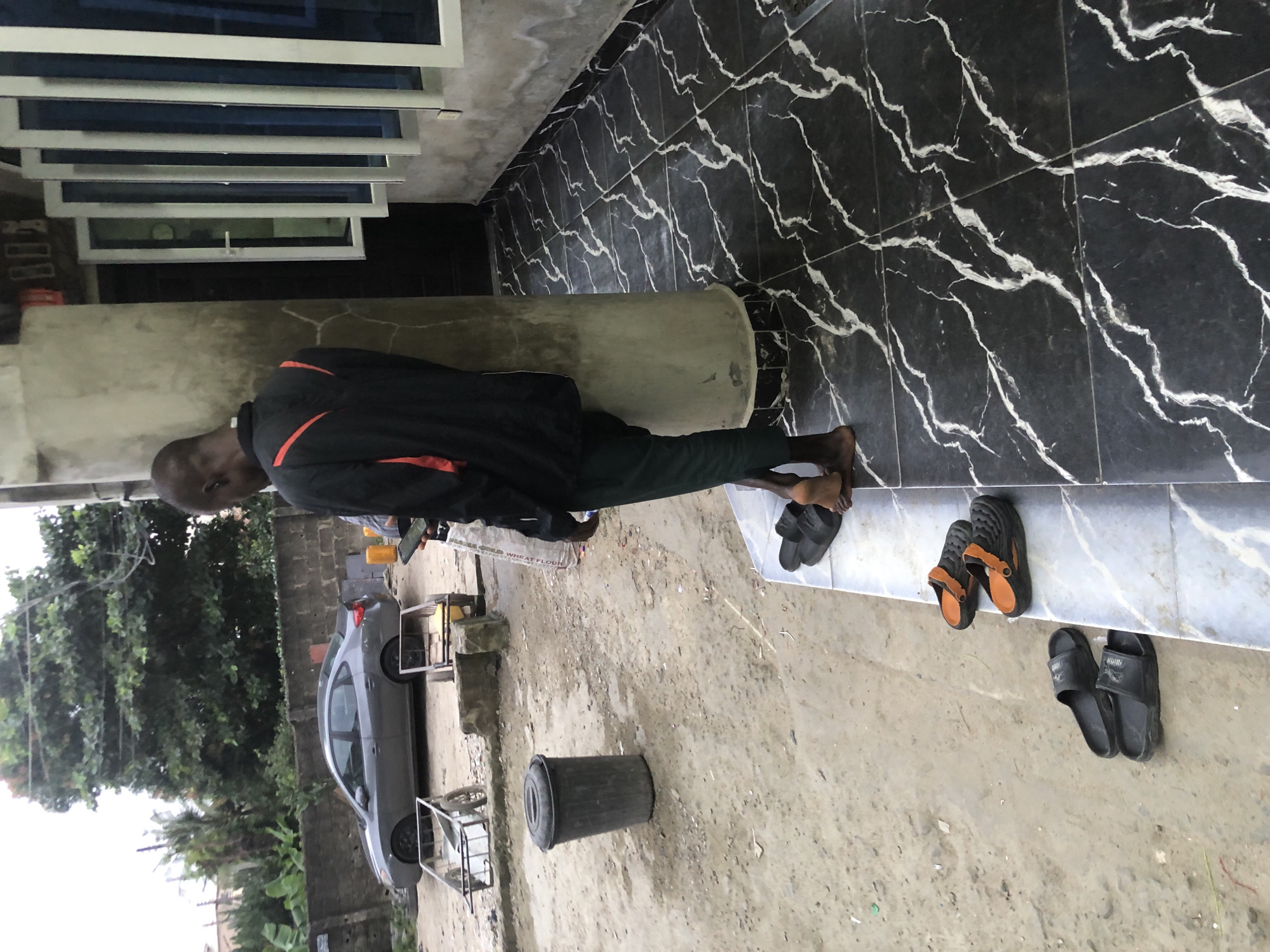 A man stands leaning against a concrete pillar, facing away from the camera. He wears a black jacket with orange accents and dark green pants. His feet are bare. To the left of the pillar, on the ground, are two pairs of black sandals and a pair of black and orange clogs. Behind the man, a building has a row of open windows with blue tinted glass. The ground is a mix of dirt and patterned black and white tiles. In the background, a grey sedan is parked on a dirt lot, and two metal carts with wheels are visible. The scene appears to be an outdoor area in Ado, Nigeria, possibly a residential or commercial space with ongoing construction or upkeep.