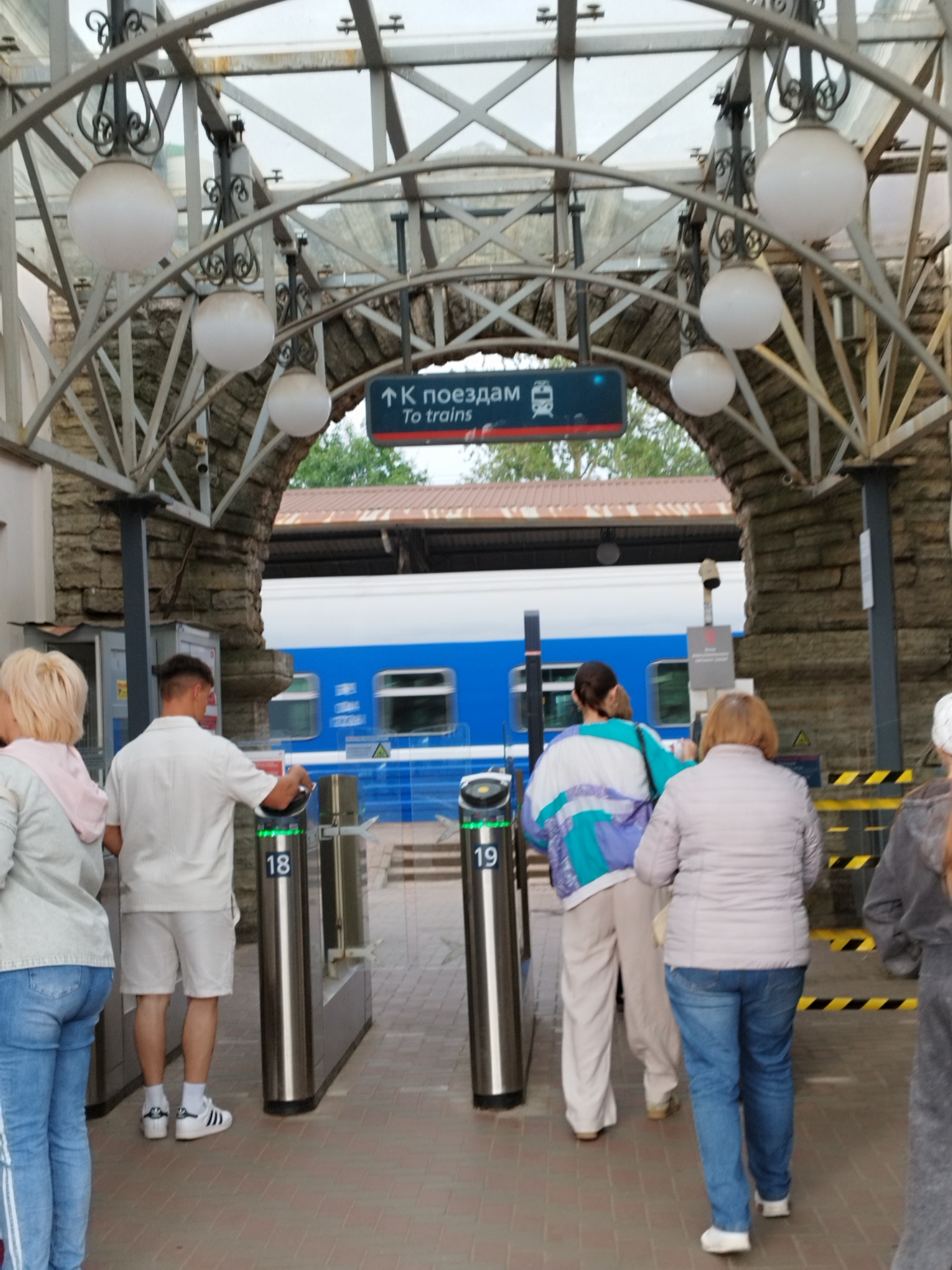 In the image, several people are seen walking through turnstiles at what appears to be a train station entrance. The turnstiles have illuminated green indicators, suggesting they are operational. One man, wearing a white shirt and shorts, is seen interacting with a turnstile. Further back, a blue and white train is visible on the tracks. The scene is under a covered walkway supported by an arched metal structure, with large spherical lights hanging from the ceiling. The walkway is lined with stone walls. The time of day appears to be daytime, and the weather seems overcast. A sign above the turnstiles reads "К поездам" and "To trains" in English, indicating directions to the trains. The overall atmosphere is one of transit and movement, with people en route to catch a train.