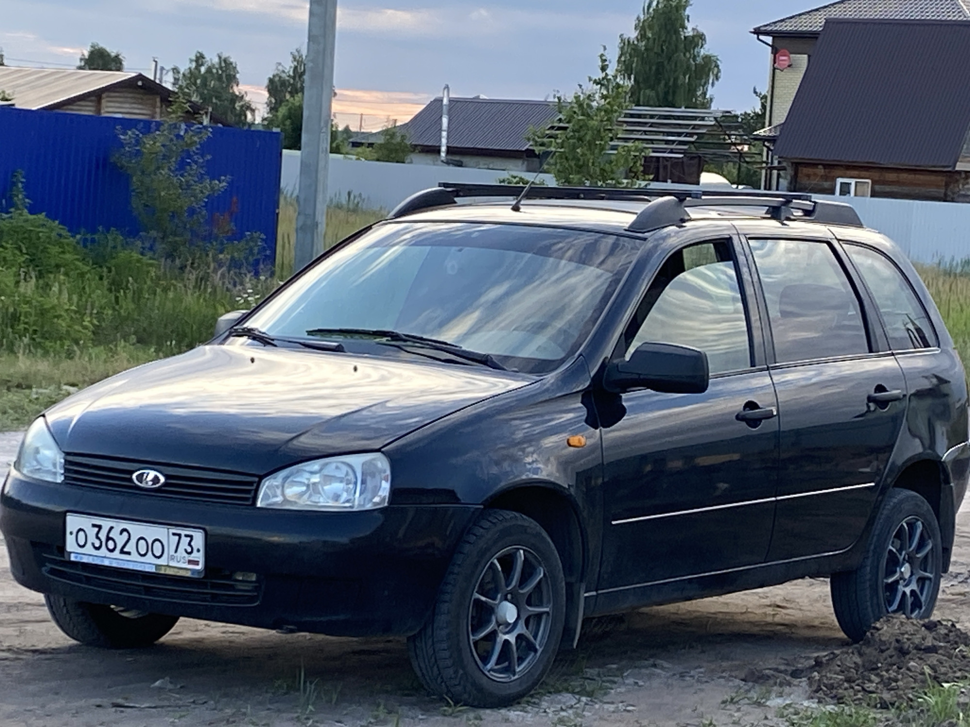 This media file shows a black Lada Kalina station wagon parked outdoors on a dirt road. The car is the primary subject, positioned in the foreground and taking up most of the frame. It has a roof rack and appears to be a model from the early 2010s. Visible text on the license plate reads "O 362 OO 73 RUS".
The scene is set in a rural or suburban environment, likely in Ulyanovsk, Russia, as indicated by the license plate's region code (73). In the background, there are several houses with dark roofs, wooden fences, and a bright blue metal fence. Trees and foliage are scattered throughout the background, suggesting a natural setting. The sky is partly cloudy, with hints of sunset colors indicating it is late afternoon or early evening. The lighting is soft, consistent with the golden hour. There are no people visible in the image. The overall impression is a quiet, everyday scene of a car parked in a residential area.