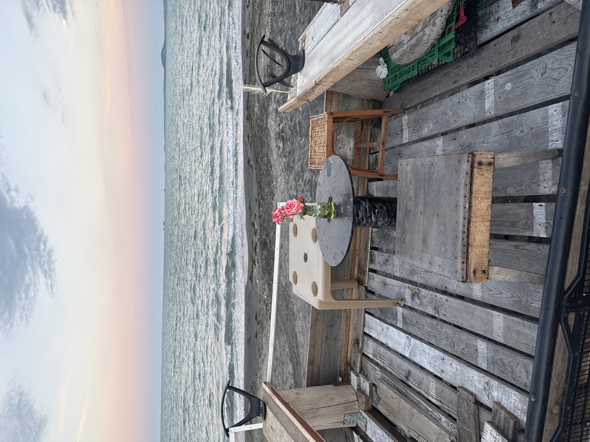 The image depicts an outdoor seating area on a wooden platform overlooking a beach and the Black Sea at dusk. The location is identified as a cafe or bar on the Burgas North Beach, Bulgaria. In the foreground, a rustic wooden deck is laid out with various pieces of salvaged wood. A small, dark circular table is set with a glass vase containing a bouquet of pink roses. To the left of the table, a tan plastic chair is placed, and to the right, a small wooden stool with a woven seat is positioned. Further to the left, a bench-like structure with a black seat is visible, made from wooden planks and metal. In the background, the sandy beach meets the calm sea, with gentle waves rolling onto the shore. The sky displays a gradient of colors, transitioning from a soft pink and orange near the horizon to pale blue and grey clouds higher up, indicating the sunset. Several distant boats or ships are faintly visible on the horizon. The overall atmosphere is tranquil and somewhat unpretentious, suggesting a casual beachfront establishment.