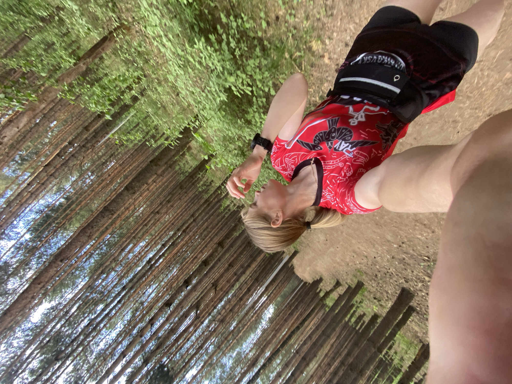A woman with blonde hair tied in a ponytail is standing on a dirt path in a forest. She is wearing a red t-shirt with a black and white design, black shorts with a black belt, and a black watch on her left wrist. The forest is dense with tall, thin trees. The photo appears to be taken from a low angle. The location is Chaikovsky, Russia.