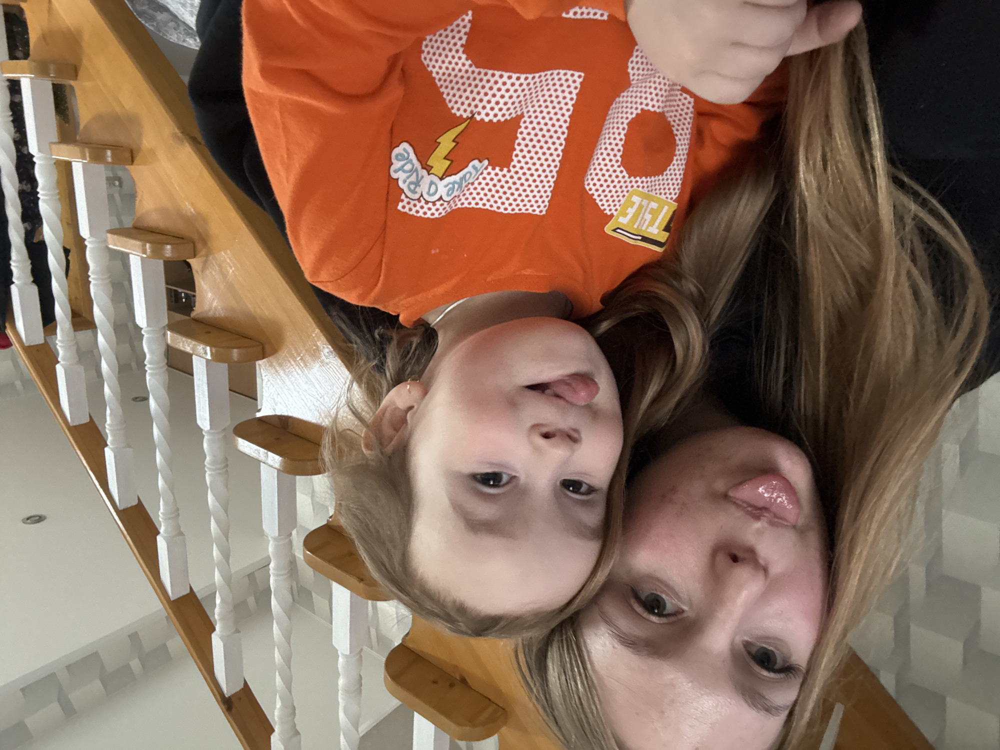 A woman and a child are lying on their backs on a staircase. They are close together, their heads aligned. Both have stuck out their tongues. The child is wearing an orange shirt with the number "50" and a lightning bolt graphic. The staircase has wooden treads and white balusters with wooden handrails. The background suggests an interior setting with white walls and possibly decorative paneling. There is no visual information to determine the location as Zhigalki, Russia.