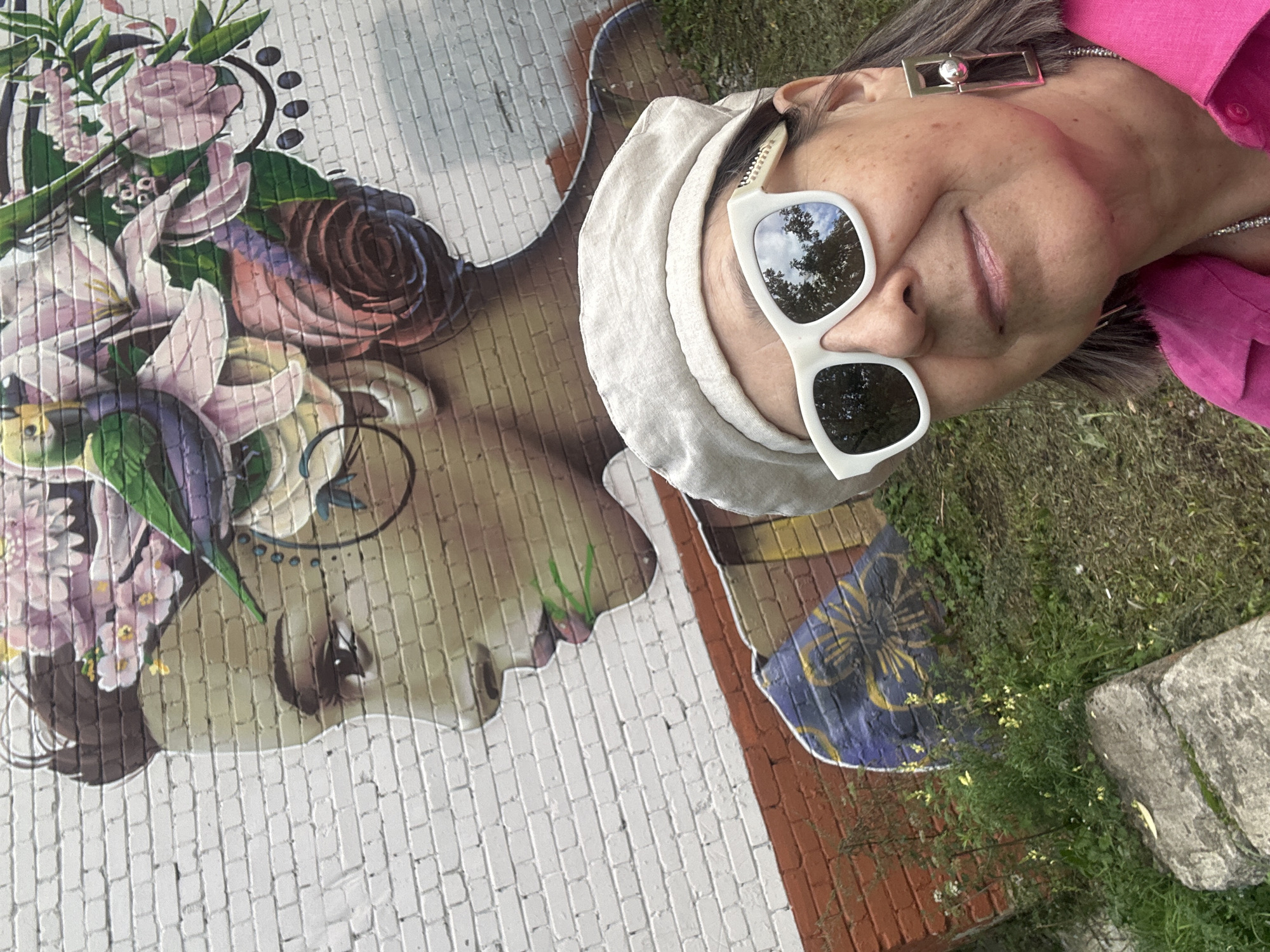 In an outdoor setting, a woman wearing a pink shirt, a light-colored beret, and white sunglasses takes a selfie in front of a large brick wall mural. The mural depicts a woman's face in profile, adorned with colorful flowers and a green bird. The lower portion of the mural shows a dark blue element with yellow floral designs. The ground in front of the mural is covered with green grass and a large gray rock. The woman's sunglasses reflect the sky and trees.